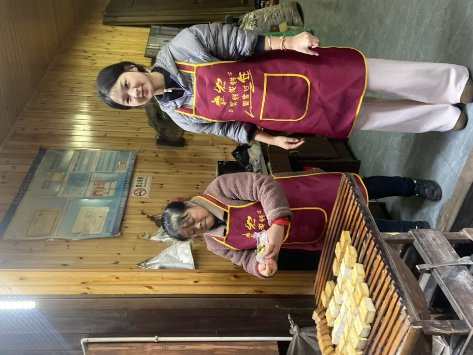 Two women wear matching maroon aprons with gold trim and Chinese characters. The younger woman stands smiling, wearing a gray puffer jacket and white pants. The older woman, in a brown sweater and dark pants, focuses on packaging small items. They stand inside a room with wooden walls and floors. A display board with documents and a "No Smoking" symbol is visible on the wall. Rows of small, square objects are displayed on a wooden rack.