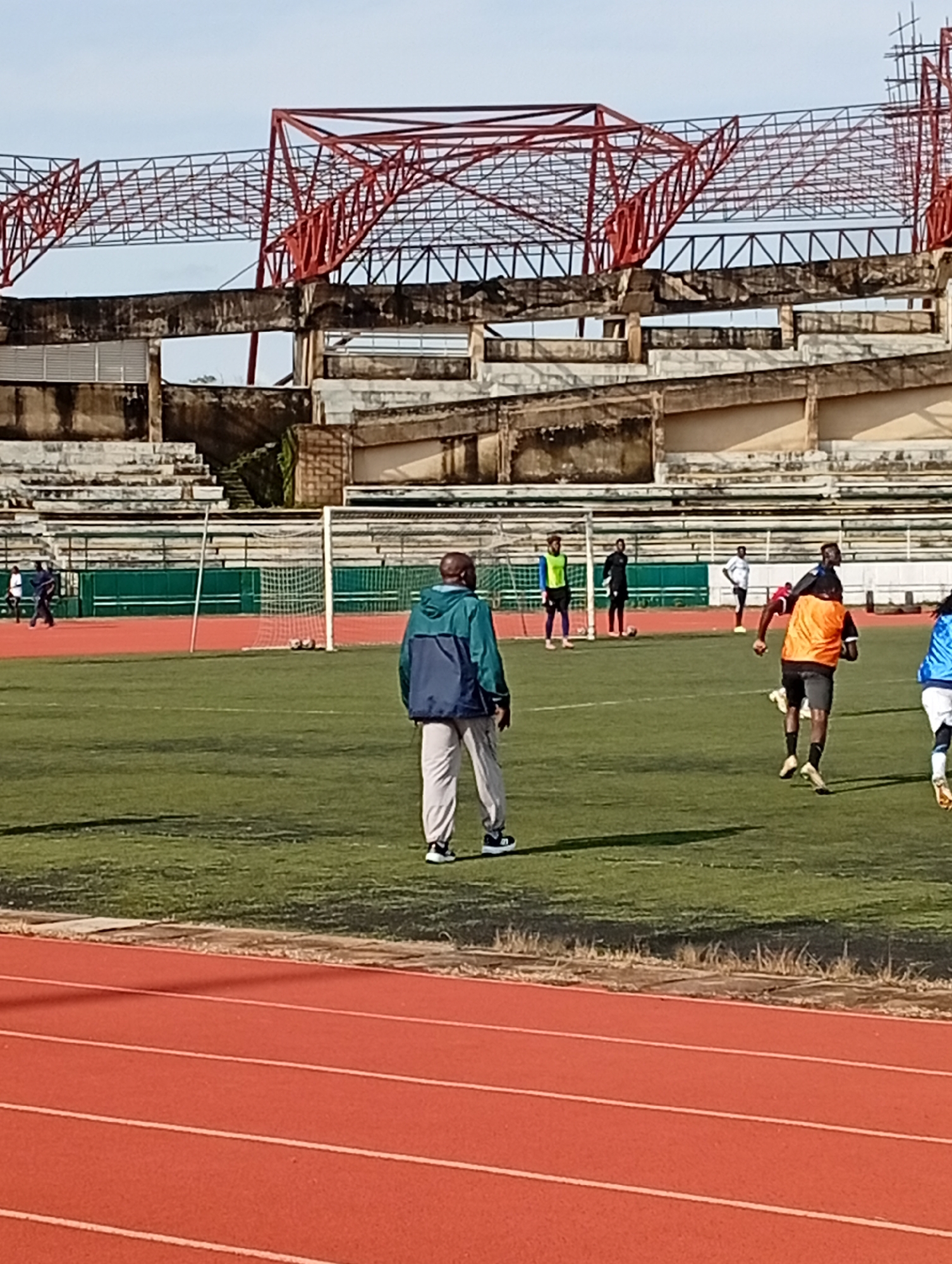 The image depicts a football training session at a stadium in New Stadium, Nigeria. In the foreground, a man wearing a blue and green jacket and light-colored pants stands facing away from the camera, observing the practice. The background shows a football field with several players engaged in training. Some players are wearing orange and blue jerseys. A soccer goal is visible on the field, and two soccer balls are on the ground near it. The stadium structure, with its red metal framework and concrete stands, is partially visible in the upper portion of the image, suggesting ongoing construction or renovation. The scene is set outdoors under a bright sky.