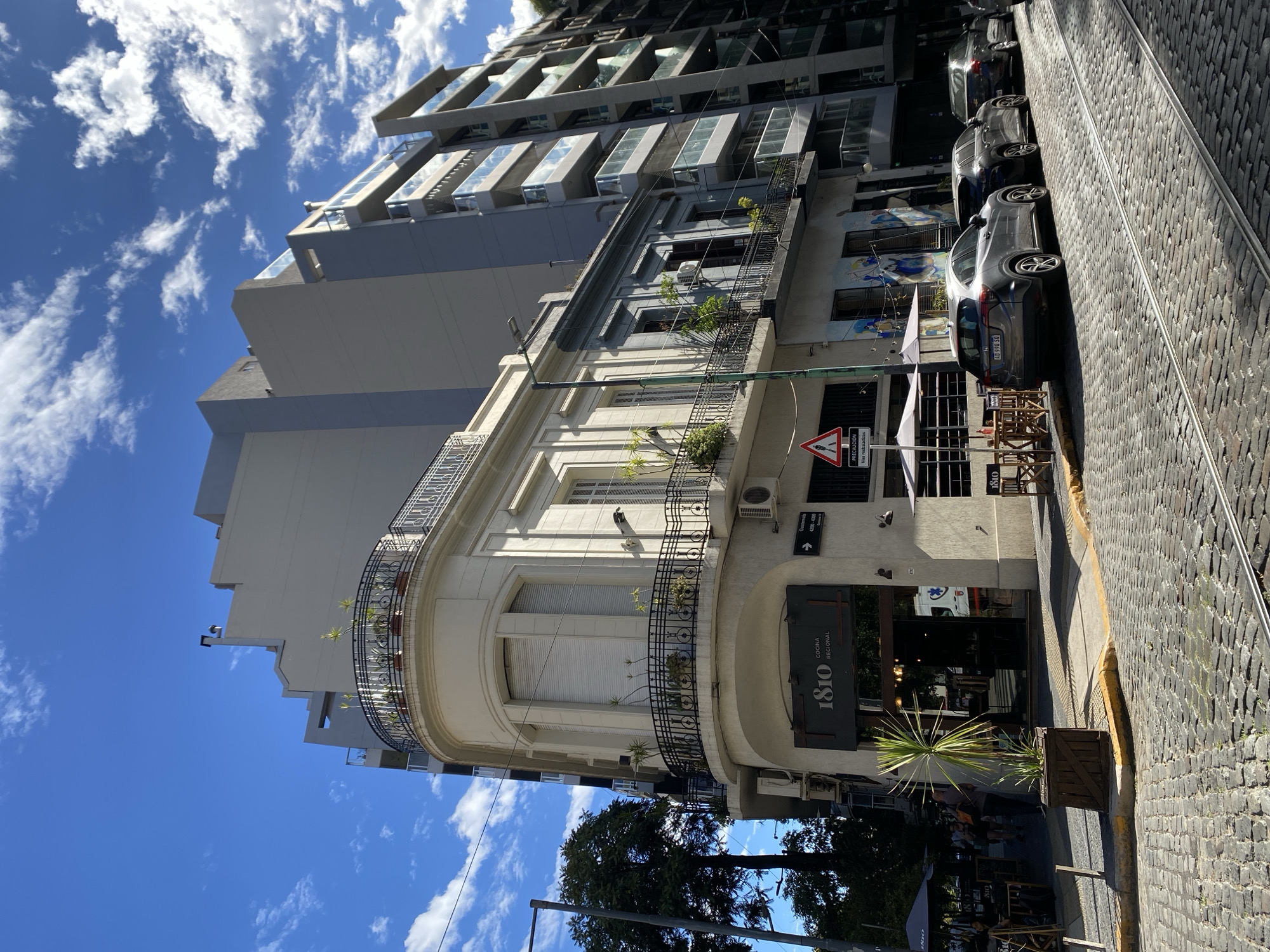The image depicts a sunlit street scene in the Palermo neighborhood of Buenos Aires, Argentina, specifically near the 4200-4300 block of Guatemala Street. The sky is bright blue with scattered white cumulus clouds.
At the focal point is a two-story, cream-colored corner building of classical architectural style, featuring ornate black wrought-iron balconies on both floors, adorned with potted plants. The ground floor operates as "1810 Cocina Regional," a restaurant with a dark entrance and an outdoor dining area furnished with wooden tables, chairs, and white umbrellas. A large potted yucca plant stands on the sidewalk in front of the establishment. Visible air conditioning units are mounted on the building's exterior.
To the immediate right of the corner building is a light blue-gray two-story building with a balcony and murals on its lower wall. Further right, a multi-story, modern apartment building with numerous glass balconies is present. To the left of the corner building, a taller, modern gray apartment building also with glass balconies rises.
The street is paved with cobblestones and features embedded metal tracks, indicative of former or current tram lines. A yellow curb delineates the sidewalk. Four passenger vehicles are parked parallel to the right curb, including a dark gray Honda HR-V (license plate AD 990 SO). An ambulance, white with red markings, is partially visible across the street, reflected in the restaurant's window. A street sign on the corner building indicates "Guatemala 4200-4300." A triangular traffic sign reading "PRECAUCION Vias resbaladizas" (Caution Slippery Roads) is visible on the right sidewalk. Multiple indistinct human figures are present on the left sidewalk, some seated at the outdoor tables, others walking. Sunlight illuminates the scene, casting distinct shadows.