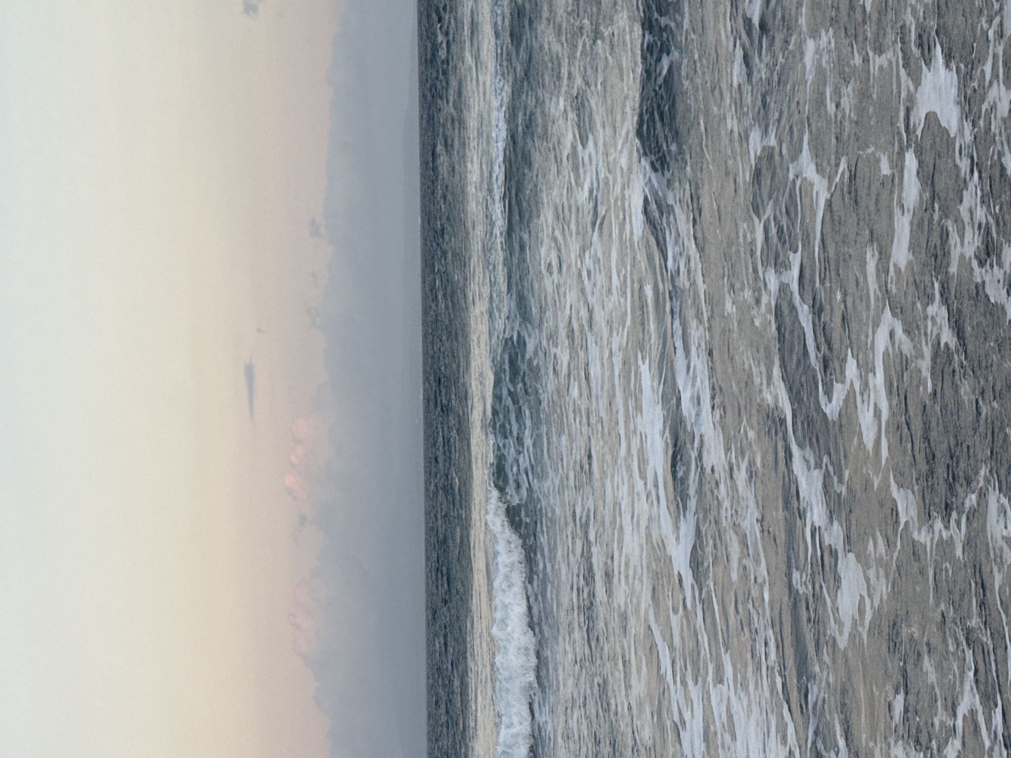 An ocean scene at dawn or dusk, with a soft pastel sky fading from pale yellow at the top to light pink and then a soft grey near the horizon. A line of indistinct clouds, tinged with pink and grey, is visible across the horizon. The sea is a deep, slightly muted blue-grey, with ripples and small waves. White foam from breaking waves is scattered across the foreground and mid-ground, creating a textured pattern on the water's surface. In the far distance, a faint, dark outline of land or mountains is visible beneath the horizon. The image appears to be taken from a low vantage point, close to the water. There are no people or man-made objects visible in the image. The scene evokes a sense of calm and natural beauty. The provided crops confirm details of the sky, clouds, and the sea with its foamy texture.