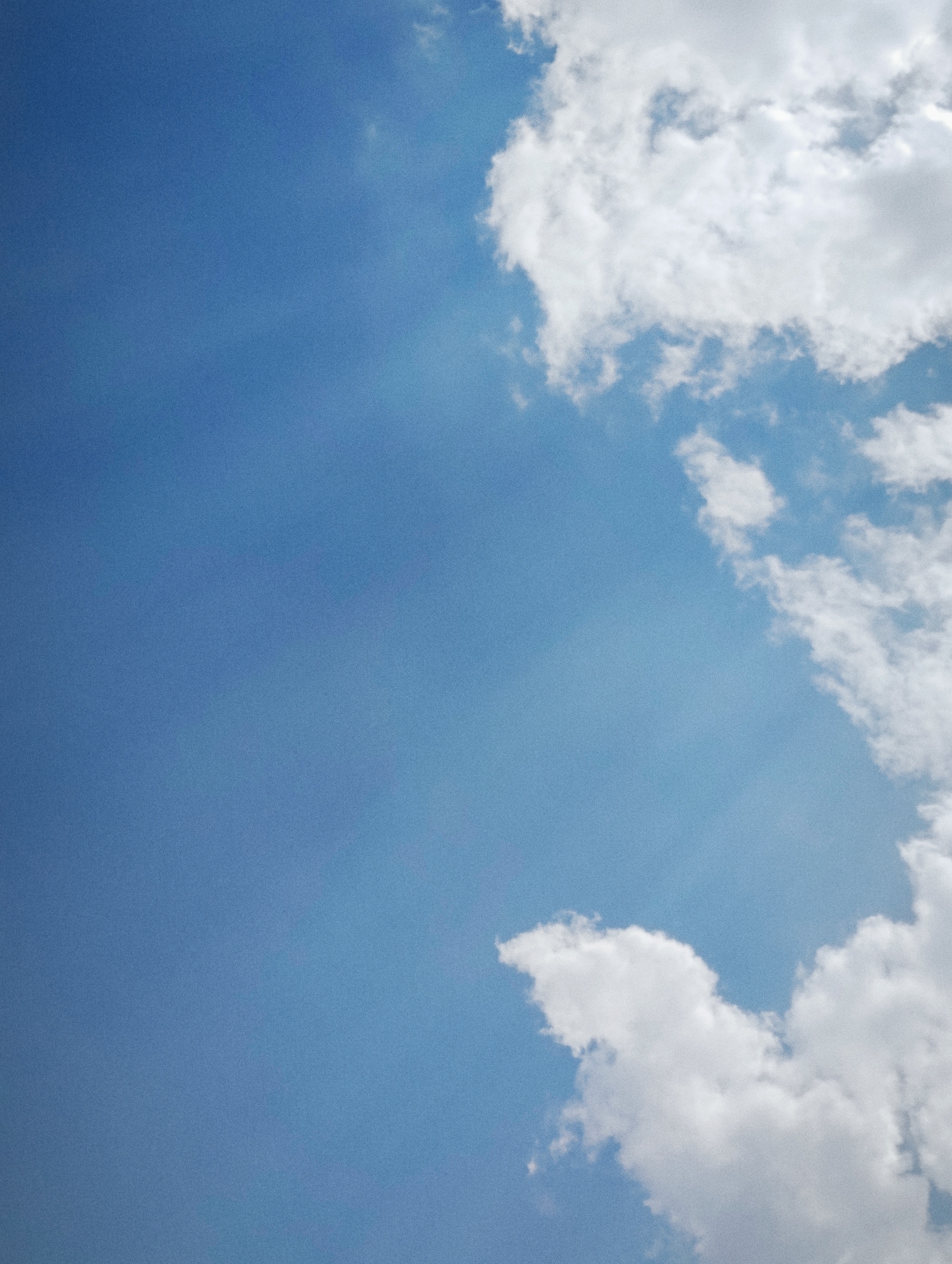 The image depicts a clear blue sky with scattered white clouds. The clouds are concentrated in the upper right and lower right portions of the frame, appearing fluffy and illuminated by sunlight. The vast expanse of the sky takes up the majority of the image. There are no people, objects, or specific landmarks visible in the scene. The environment is an outdoor, natural setting, likely a daytime view looking upwards. The image does not provide any contextual information or clues to suggest it is located in Manali, India, or any other specific city.