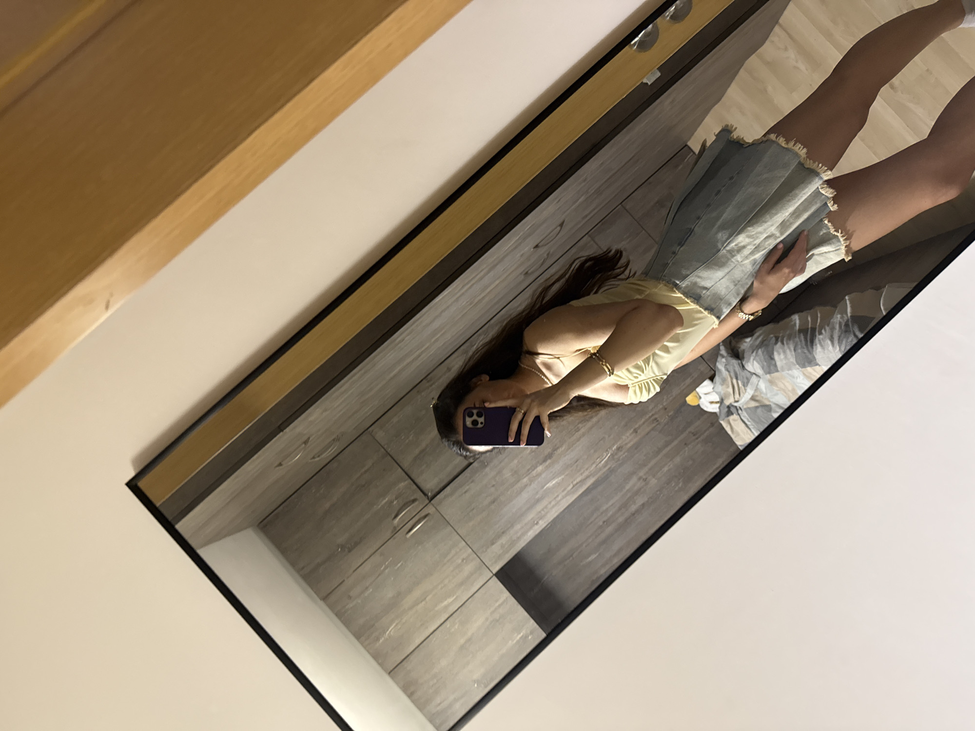 A full-length mirror reflects a woman taking a selfie with a purple smartphone. The woman has long dark hair and wears a yellow top and a denim skirt. She is standing on what appears to be a wooden floor, which is visible in the mirror's reflection. The background includes a grey cabinet or wall paneling. The mirror is framed in black and is hung on a light-colored wall. The surrounding environment is not identifiable as Plovdiv, Bulgaria.