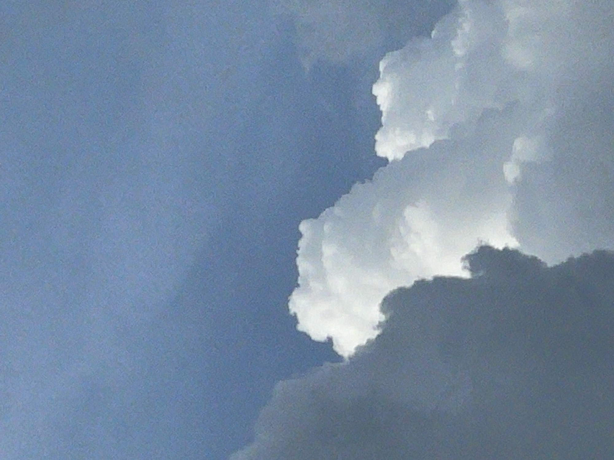 The image shows a partly cloudy sky. The left portion of the image displays a blue sky, while the right portion features a large, billowing cloud formation. The lower section of this cloud appears dark gray, gradually transitioning to white at the top. The image was taken in Nkpor, Nigeria. There are no people or interactions visible in this image.