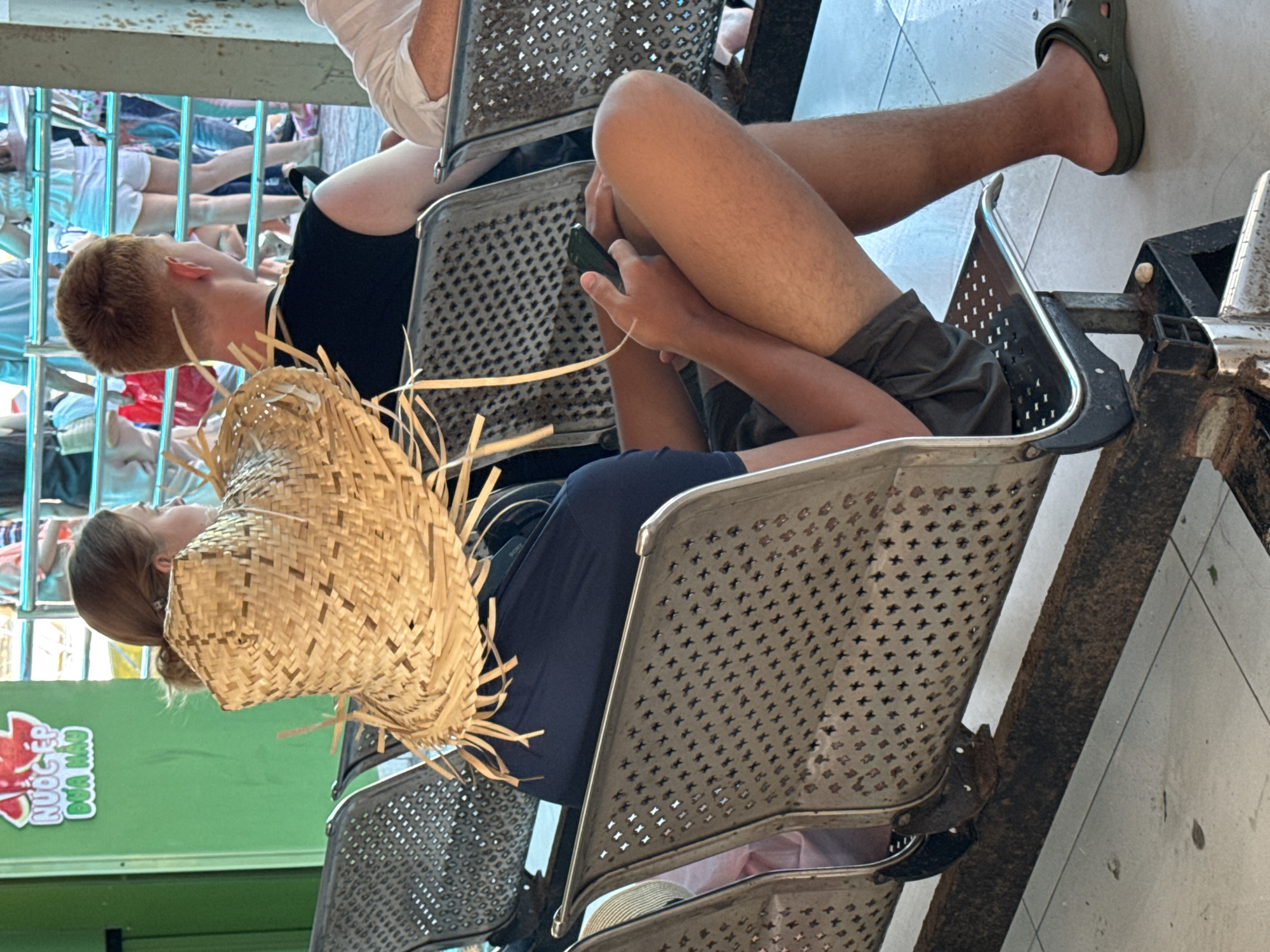 The image shows a group of people sitting on metal benches. The background is a green wall with Vietnamese text. A young man with red hair is in the foreground, wearing a black t-shirt and shorts. He is holding a mobile phone. To his left, a woman wearing a straw hat is also seated. In the background, other individuals are visible, including some wearing white shorts and t-shirts. The setting appears to be an outdoor waiting area or a public space.