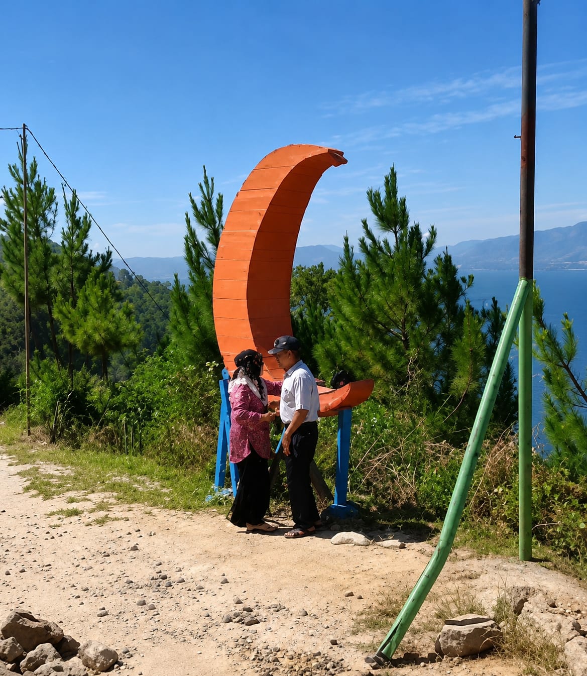 In the city of Parik Sabungan, Indonesia, an elderly couple stands on a dirt path near a large, orange, crescent-shaped sculpture. The sculpture is supported by two blue legs. The woman, wearing a pink blouse and black skirt, faces the man, who is dressed in a white collared shirt, black pants, and a baseball cap. They appear to be talking. Behind them, pine trees and lush green foliage cover a hillside. In the distance, a wide body of blue water, likely a lake, is visible, with mountains on the horizon under a clear blue sky. A tall, dark pole with a green support is positioned in the foreground to the right of the couple.