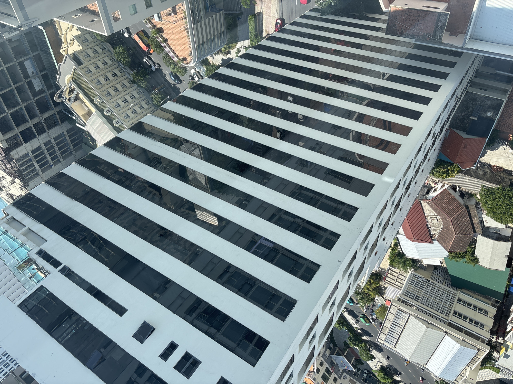 The image provides an aerial perspective of a bustling city scene in Nha Trang Ward, Vietnam, looking down from a high vantage point. The foreground is dominated by the exterior of a tall, modern white building with numerous dark, rectangular windows arranged in diagonal stripes. Below and to the right, a complex urban landscape unfolds, featuring a variety of buildings with different architectural styles and roof types, including red-tiled roofs and metallic grey roofing.
Several streets are visible, with traffic including cars, motorcycles, and a yellow bus navigating the roads. Trees and green spaces are interspersed among the buildings, adding a touch of nature to the urban environment. In the distance, other tall buildings and construction sites are present, indicating ongoing development within the city. On the top of one of the buildings in the foreground, a swimming pool with turquoise water and a person near it can be seen. The overall impression is one of a dense and active city, captured from an elevated and oblique angle.