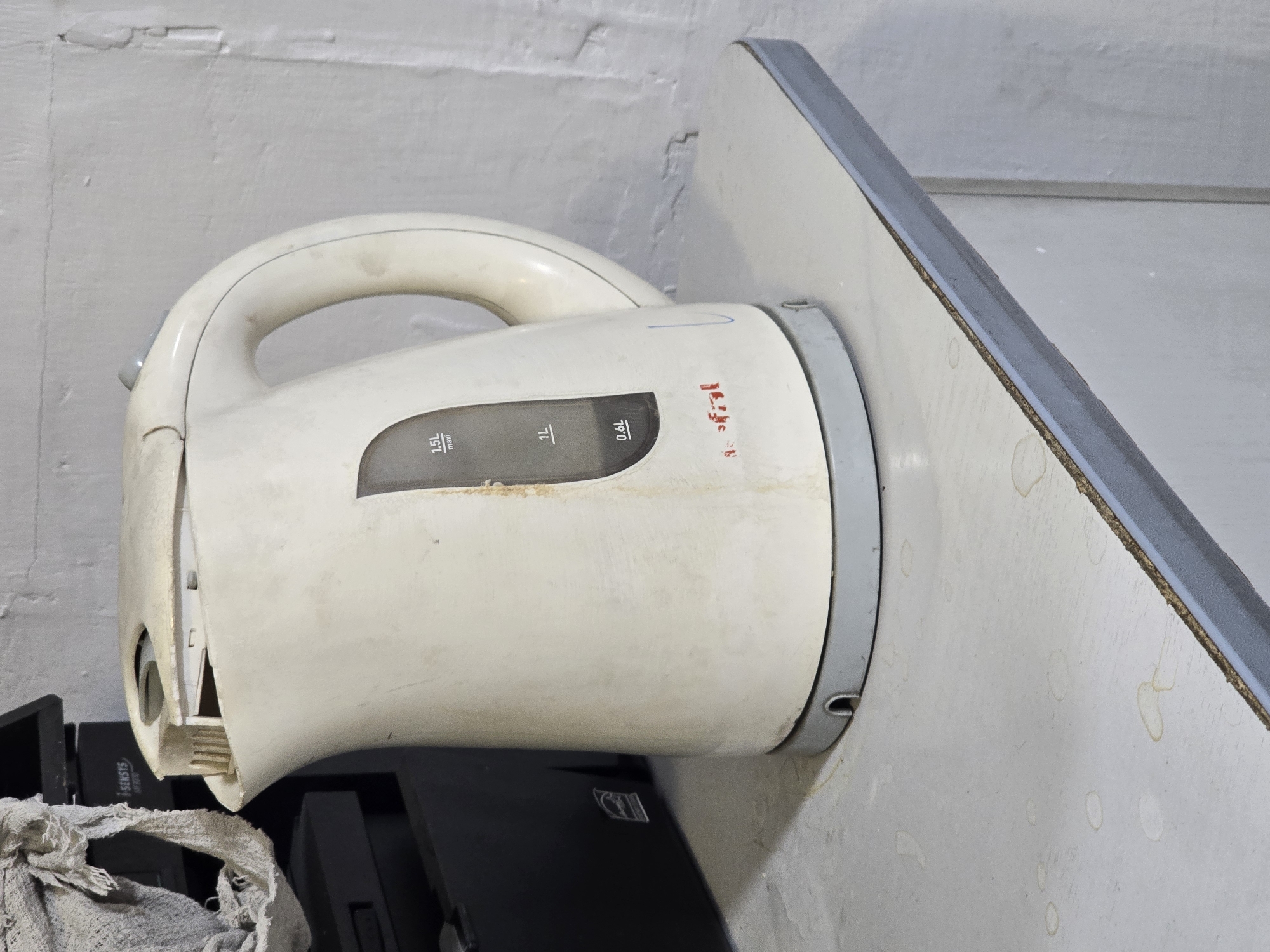 The image shows a dirty, white electric kettle with a visible water level indicator marked in liters. The kettle is placed on a worn, light-colored countertop that has some liquid stains. In the background, a dark-colored object, possibly a printer or scanner, is partially visible, with a crumpled piece of light-colored fabric draped over it. The wall behind the objects appears to be textured and painted white, with some imperfections. The scene is indoors, likely in a domestic or utilitarian setting, and does not provide any specific indicators to place it in Ulan-Ude, Russia, or any other city. There are no people, actions, or interactions depicted in the image.