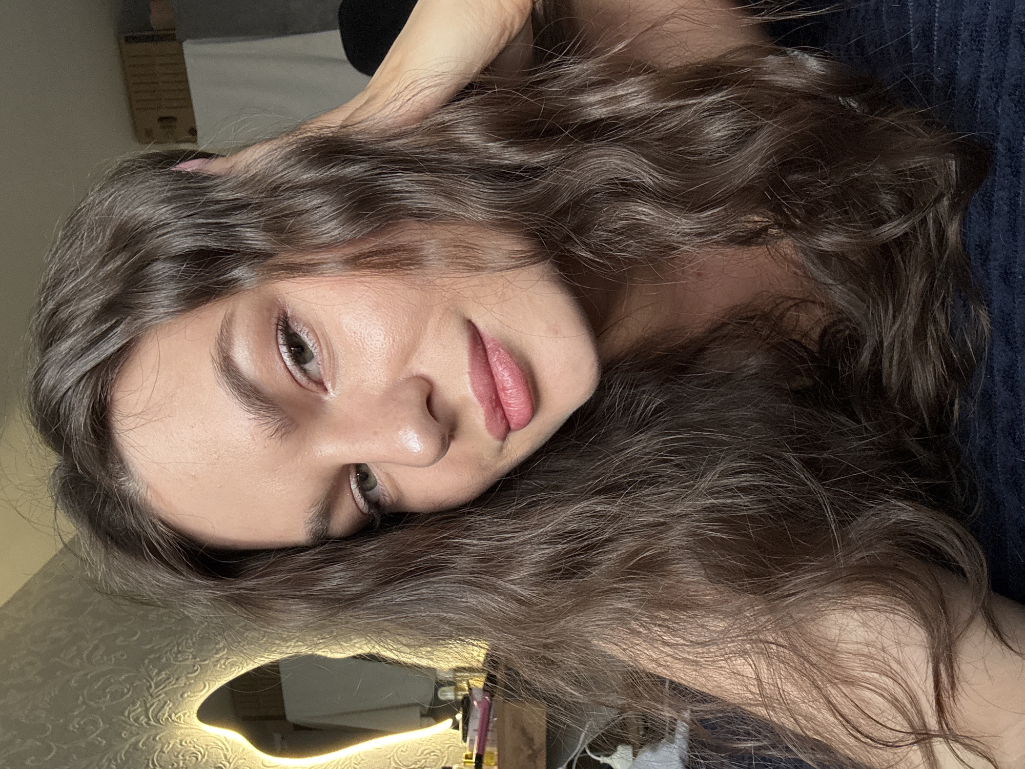A young woman with long, wavy brown hair is lying down, looking into the camera. She is wearing makeup, including pink lipstick and eyeshadow. Her right hand is resting on the back of her head, and her left arm is visible across the lower part of the frame. The background is blurred but appears to be an indoor setting with a textured wallpaper and a lit mirror. There is a dark blue fabric underneath her. The scene is a close-up shot, focusing on her face and upper body. There is no identifiable indication of location in the city of Chaikovsky, Russia.