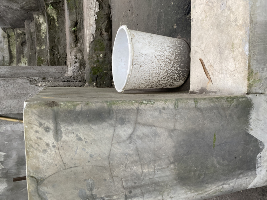 The image shows a white plastic bucket sitting on a concrete surface. The bucket is positioned between two concrete walls. The bucket has a weathered, dirty appearance. The surface the bucket sits on appears to be part of a set of concrete steps that extend upward and out of the frame. There is some green moss growing on the steps. The scene appears to be in an outdoor location. There are no people in the scene. The location is in Kecamatan Piyungan, Indonesia, at latitude -7.825 and longitude 110.453.