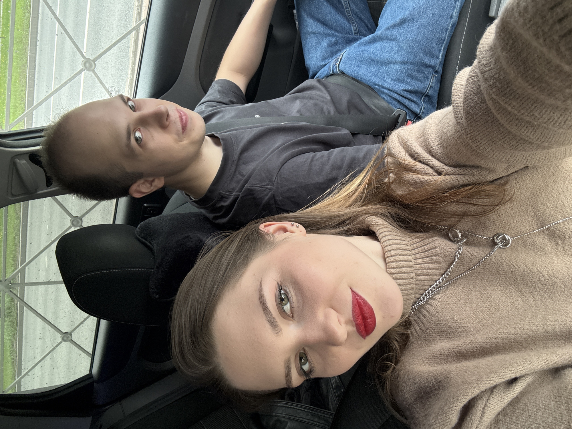 Two individuals are in the backseat of a car. The individual in the foreground has long brown hair, fair skin, green eyes, and is wearing red lipstick and a beige sweater, along with a silver necklace. They appear to be the one taking the photograph. The second individual is in the background, with fair skin, short hair, a grey t-shirt, and blue jeans. Both individuals have their seatbelts on and are facing the camera. The scene is located in a car with black interior. Outside the car window, it appears to be raining, and there is a view of a structure resembling a fence or barrier. Given the architectural style visible outside the window, the location is potentially Saint Petersburg, Russia.