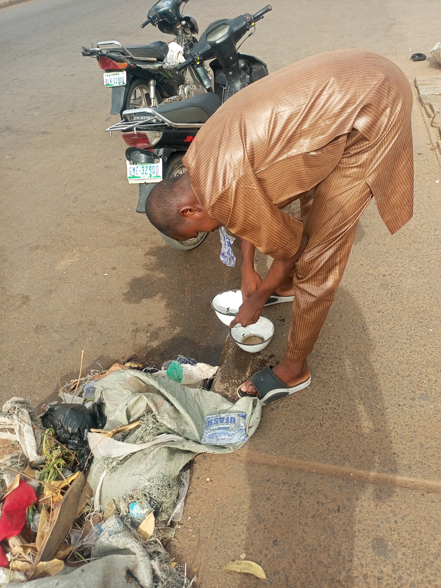 A man wearing a brown, striped robe and dark sandals is bending over a pile of trash on a street in Nigeria. He is holding two white bowls that appear to contain a liquid, possibly water. Two motorcycles are parked behind him, their license plates reading "BORNO BLR-119UU" and "GME-32900". The trash pile contains various items including a black plastic bag, pieces of fabric, and a package labeled "UFASH". The location is Tempure, Nigeria.