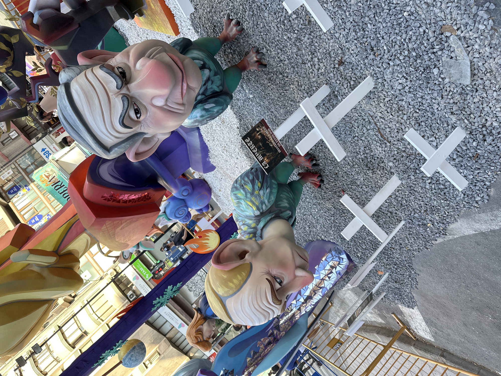 This image captures a scene in Alicante, Spain, featuring large, colorful, caricature-like figures that appear to be part of a street festival or display, possibly a "Falla" celebration, given the context of Alicante. The figures are exaggerated and whimsical, with one depicting an older man with grey hair and a stern expression, and another a creature-like figure with large ears and a smiling, slightly mischievous face. They are made of vibrant materials, with detailed sculpting and painting, suggesting they are handcrafted works of art.
The figures are positioned outdoors, set against a backdrop that includes buildings with visible storefronts and signage, indicating an urban setting. The ground is covered in gravel and asphalt, with several white cross-like structures placed around the figures, possibly as part of the display or for crowd control. In the background, glimpses of people can be seen, suggesting the presence of an audience or participants in the event.
A notable detail is a small, black sign held by one of the figures, which contains text in what appears to be Spanish. The text is difficult to read clearly in the main image but appears to be "29 MARZO INVITAN A LA MESA DE ESTOS DOS". This suggests a specific date, "March 29th," and an invitation to "the table of these two," possibly referencing the figures or a related event. The overall atmosphere seems festive and lively, typical of a Spanish cultural celebration. The lighting suggests it might be daytime, with overcast conditions or shadows from surrounding structures.