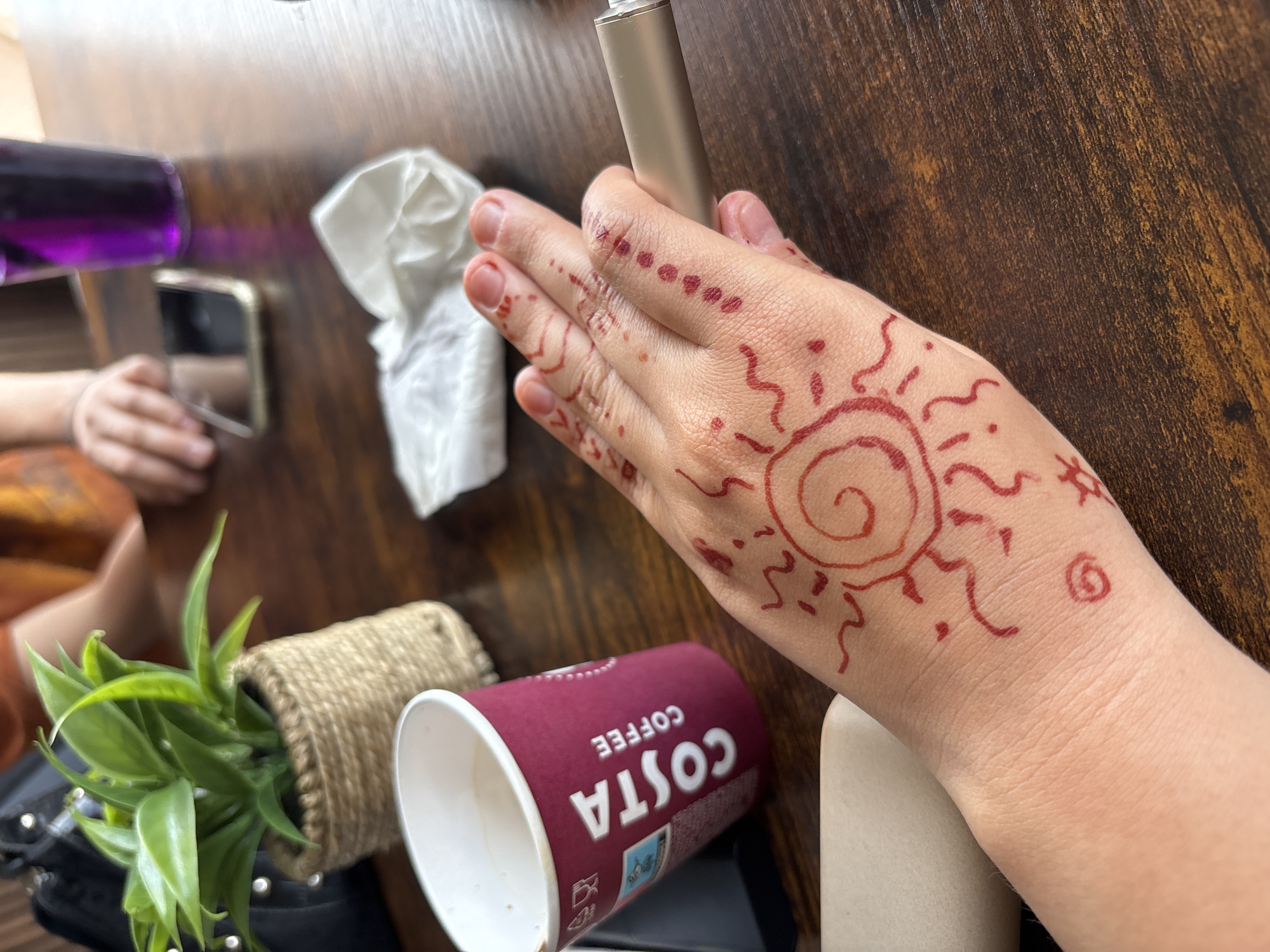 A close-up shot shows a person's hand with henna art on a wooden table. The henna design includes a sun with radiating lines and dots along the fingers. A Costa Coffee cup is visible to the left of the hand, and a metallic object, possibly a vape pen, is being held by the hand. In the background, a small potted plant, a phone, and a glass of purple liquid are present. The overall setting appears to be a casual indoor environment, likely a cafe or a home. There is no information in the image to suggest the location is Garmen, Bulgaria.