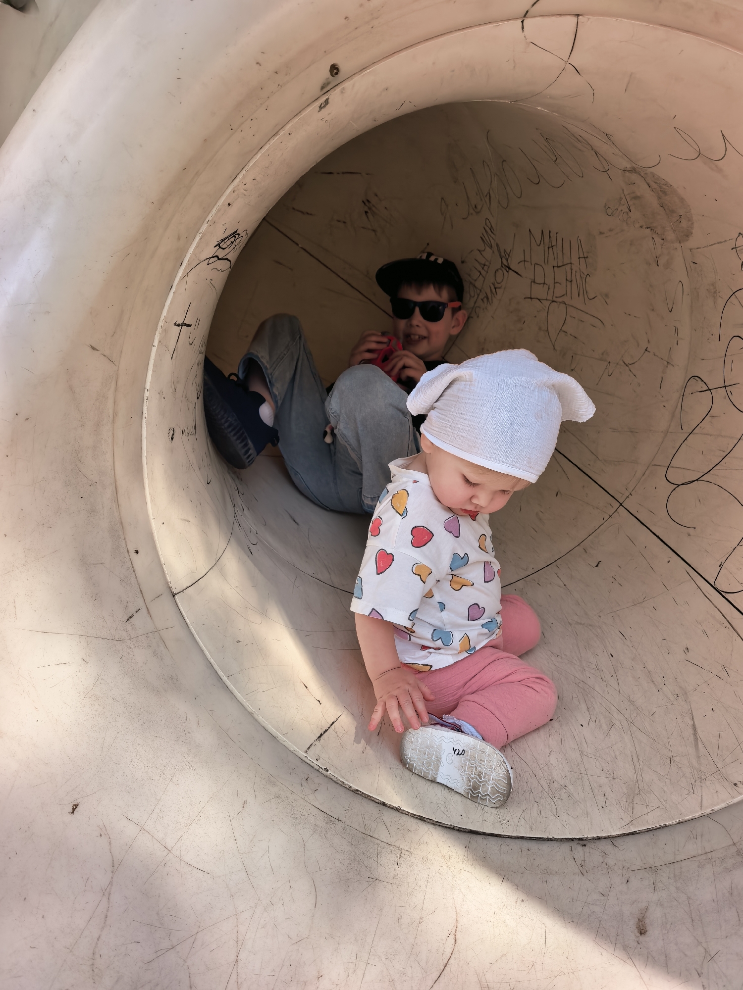 Two children are inside a large, curved, white plastic tube, likely part of a playground slide. The child in the foreground is a toddler, wearing a white hat with ear-like protrusions, a white t-shirt with colorful heart patterns, and pink pants. The toddler is sitting and looking downwards. In the background, a slightly older child wearing sunglasses and a black baseball cap is lying down, holding a red object. Both children appear to be indoors or in a shaded area, as indicated by the lighting and shadows within the tube. The interior of the tube is marked with dark scribbled graffiti. The image does not provide any specific details that would allow for localization within the city of Sterlitamak, Russia.