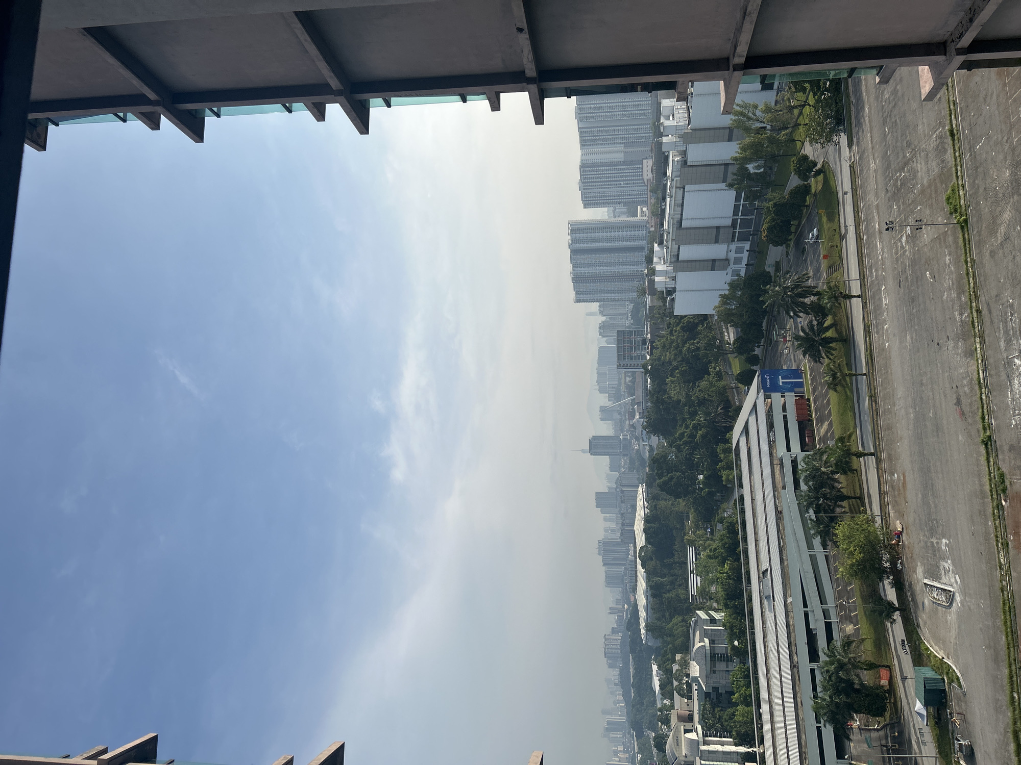 The image shows a high-angle, eye-level view of a cityscape, likely taken from a high-rise building in Kuala Lumpur, Malaysia. The foreground is partially framed by the edge of a concrete structure, possibly a balcony or the roof of the building from which the picture was taken. The sky is a gradient of blue, fading into a hazy, whitish-gray towards the horizon. Below the sky is a dense urban area, comprising a mix of high-rise buildings, green spaces (trees, parks), and streets. The buildings range in size and design, with some appearing modern and others possibly older. In the lower right portion of the frame is a large paved area resembling a parking lot or open ground with street lights. Adjacent to the paved area is a road. The image exhibits a slightly rotated orientation, with the urban grid appearing tilted. The overall impression is one of dense urban development under a hazy sky.
