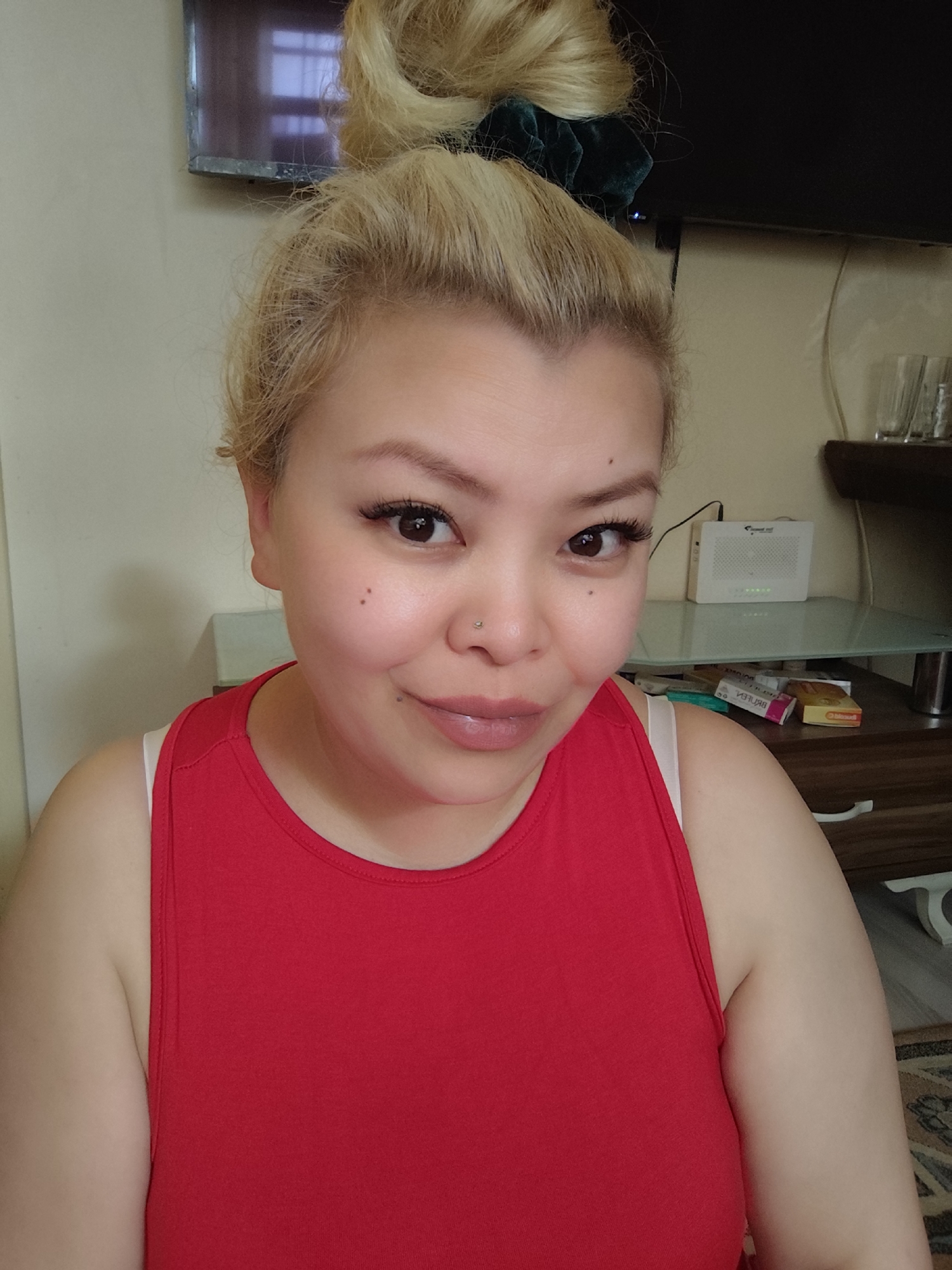 A woman with blonde hair styled in a bun secured with a dark green scrunchie is posing for a selfie. She is wearing a red tank top. She has a mole on her left cheek and a nose piercing in her left nostril. The background includes a TV mounted on the wall, a white router on a glass-topped table, glasses, and some medication boxes. The location is implied to be in Sarıyer, Turkey.