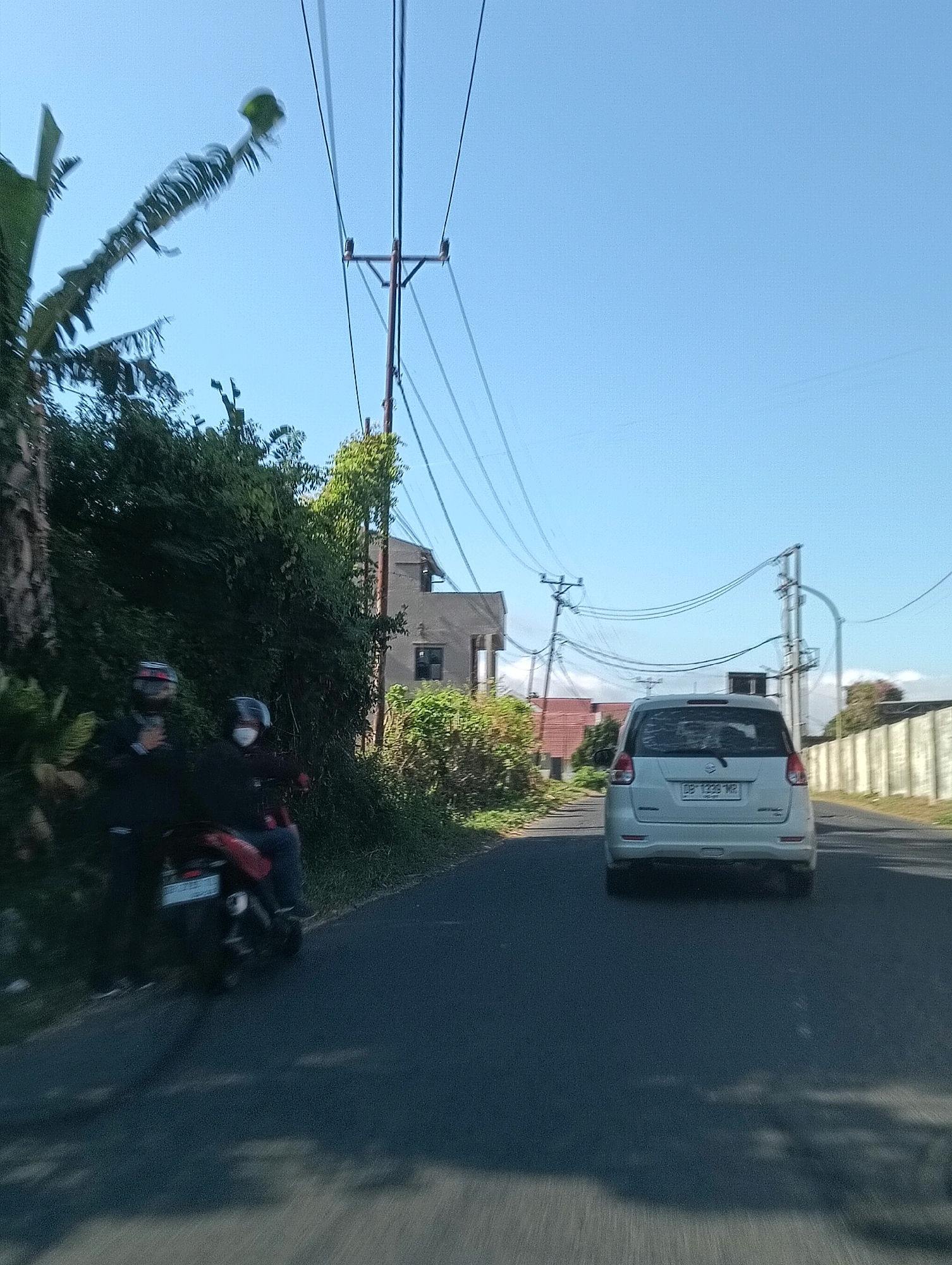 A white, four-door sedan is traveling on a paved road. The vehicle has a license plate that reads "DB 1339 MR". On the left side of the road, two individuals are riding a motorcycle. The motorcycle has a red seat and a license plate with some characters on it. The rider in the front is wearing a dark jacket and a helmet. The rider in the back is wearing a dark jacket, a helmet, and a white face mask. The road is lined with lush green vegetation and trees. In the background, there are several buildings, including a multi-story structure with a dark facade. Overhead, power lines are strung between poles. The sky is clear and blue, indicating a sunny day. The scene appears to be in an urban or suburban area of Tomohon, Indonesia, characterized by its tropical foliage and modest architecture.
