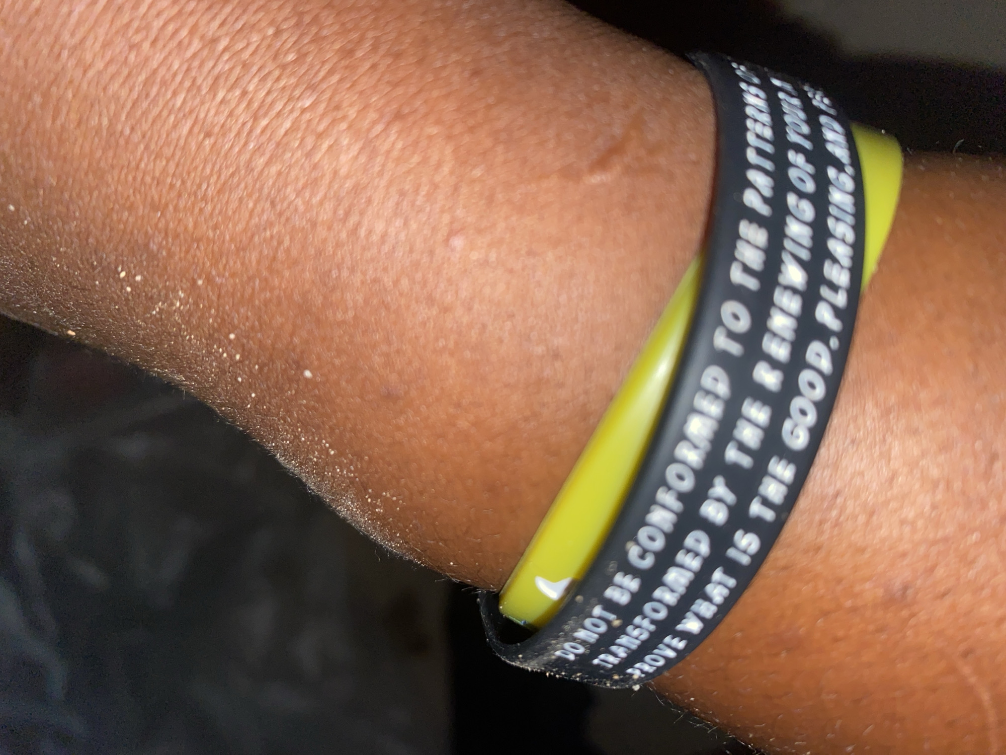 The image shows a close-up of a person's arm wearing a black and yellow wristband. The wristband has text printed on it in white letters. The text reads, "DO NOT BE CONFORMED TO THE PATTERNS OF THIS WORLD, BUT BE TRANSFORMED BY THE RENEWING OF YOUR MIND. THEN YOU WILL BE ABLE TO TEST AND APPROVE WHAT IS THE GOOD, PLEASING AND PERFECT WILL OF GOD." The arm appears to be tanned and has some texture visible. The background is dark and out of focus. The location of Jauro Yinu, Nigeria is not discernible from the image.