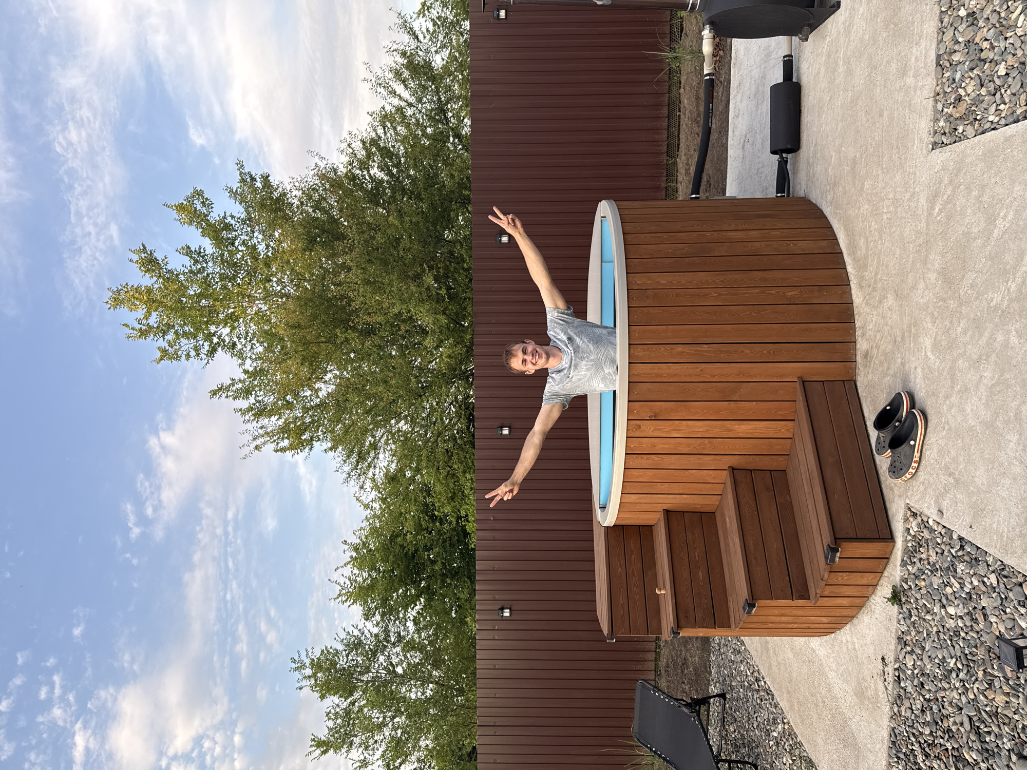 The image shows a man standing in a wooden hot tub with blue water. He is smiling and holding up two fingers in a peace sign. He is wearing a grey t-shirt. The hot tub is surrounded by wooden decking and steps. To the left of the hot tub is a brown fence and a large green tree. To the right of the hot tub is a concrete patio with gravel and a black lounge chair. There are a pair of black crocs with red stripes on the patio. The sky is blue with some white clouds. The time of day appears to be late afternoon or early evening. The weather is clear and sunny. The location is Starokorsunskaya, Russia.