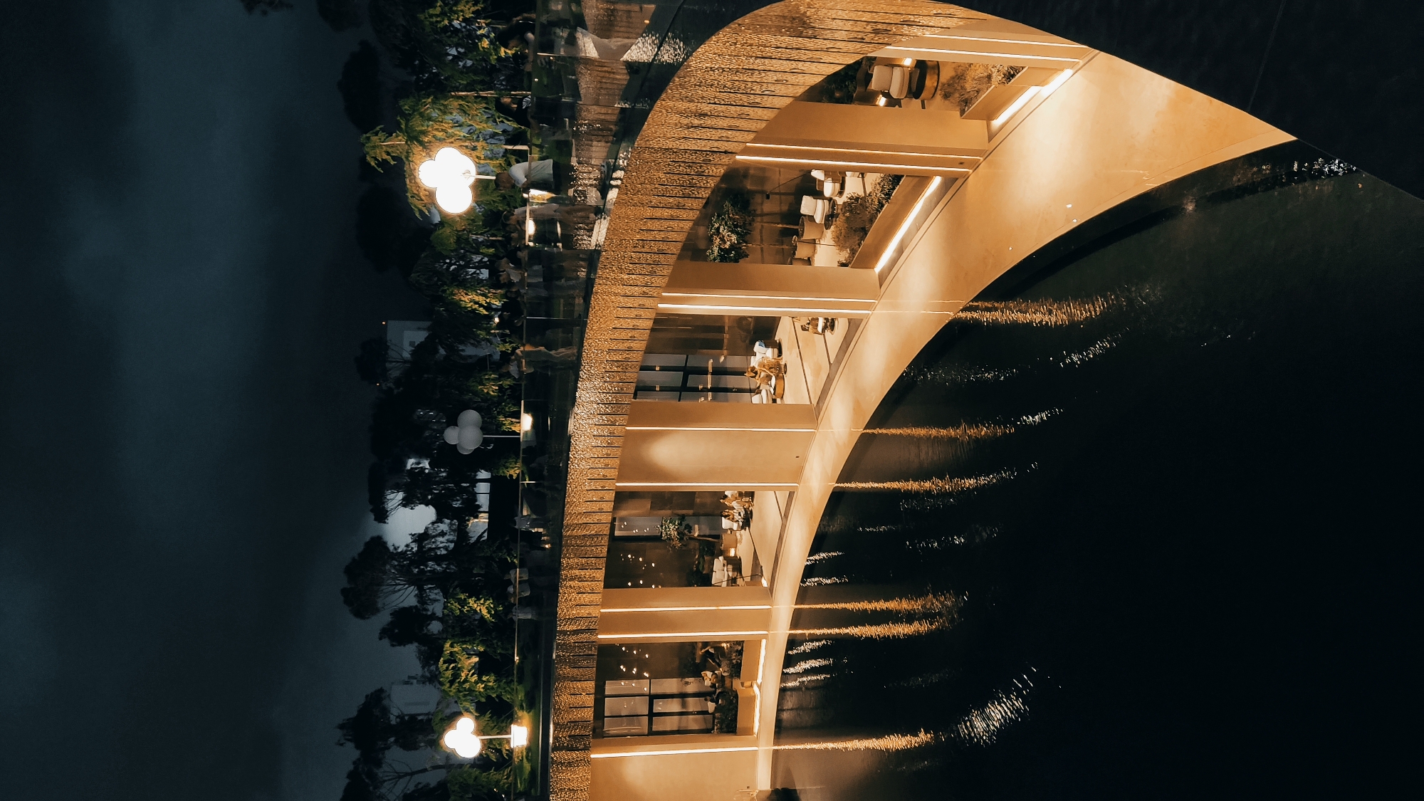 An aerial, wide-angle shot depicts a curved, modern building with large glass windows and illuminated interiors at night. The structure is situated alongside a dark, reflective body of water, possibly a pool or canal, which mirrors the building's warm interior lights. People are visible walking along a balcony or elevated walkway above the water and behind a decorative, patterned railing. The background features trees and foliage, also illuminated by decorative lamps shaped like clouds. In the distance, the dark night sky is visible, with some clouds. The overall impression is one of an upscale, contemporary establishment, likely a restaurant or lounge, in an urban setting. Based on the architecture and outdoor elements, this location could be the "Galaktika" Entertainment Center in Krasnodar, Russia, known for its unique design and nighttime ambiance.