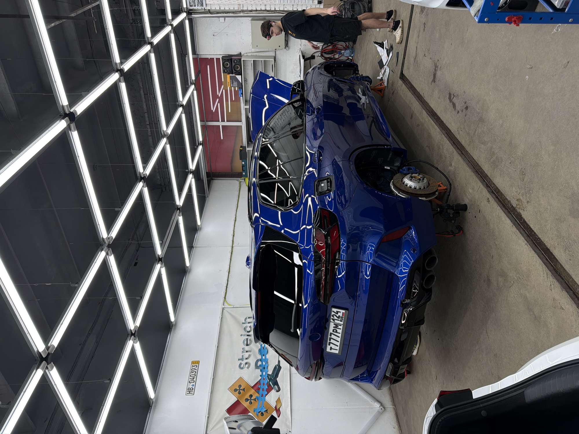 The scene is an indoor auto repair shop. A blue SUV with its hood open is being worked on by a person standing beside it. The person appears to be focused on the vehicle and is wearing a black shirt, dark shorts, a cap, and white/beige shoes. One wheel is removed from the car with the brake assembly exposed. Repair equipment is nearby. The shop is well lit with a grid of overhead lights. A banner with "stretch" printed on it is visible on the wall. A license plate on the vehicle reads "T777PM 124," which indicates the vehicle is registered in Krasnoyarsk, Russia.