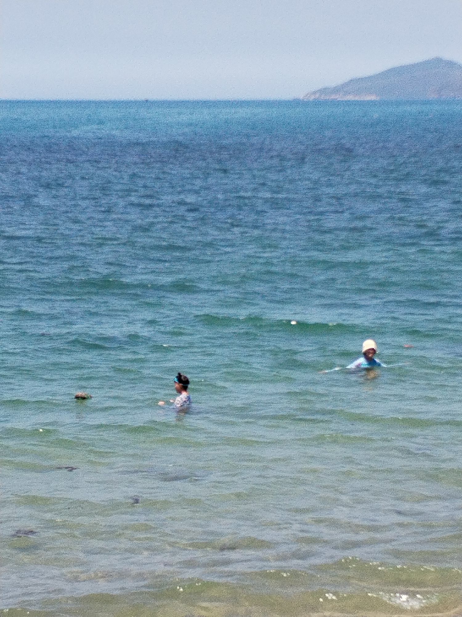 The image depicts two people swimming in the ocean near the coast of Bac Nha Trang Ward, Vietnam. The water is blue and slightly choppy. One swimmer is closer to the shore, wearing dark goggles and a patterned bathing suit. The other swimmer is further out, wearing a light-colored hat and a blue shirt. In the background, a distant landmass is visible on the horizon. The sky is clear and bright. The bottom of the image shows shallow water with some rocks or debris visible.