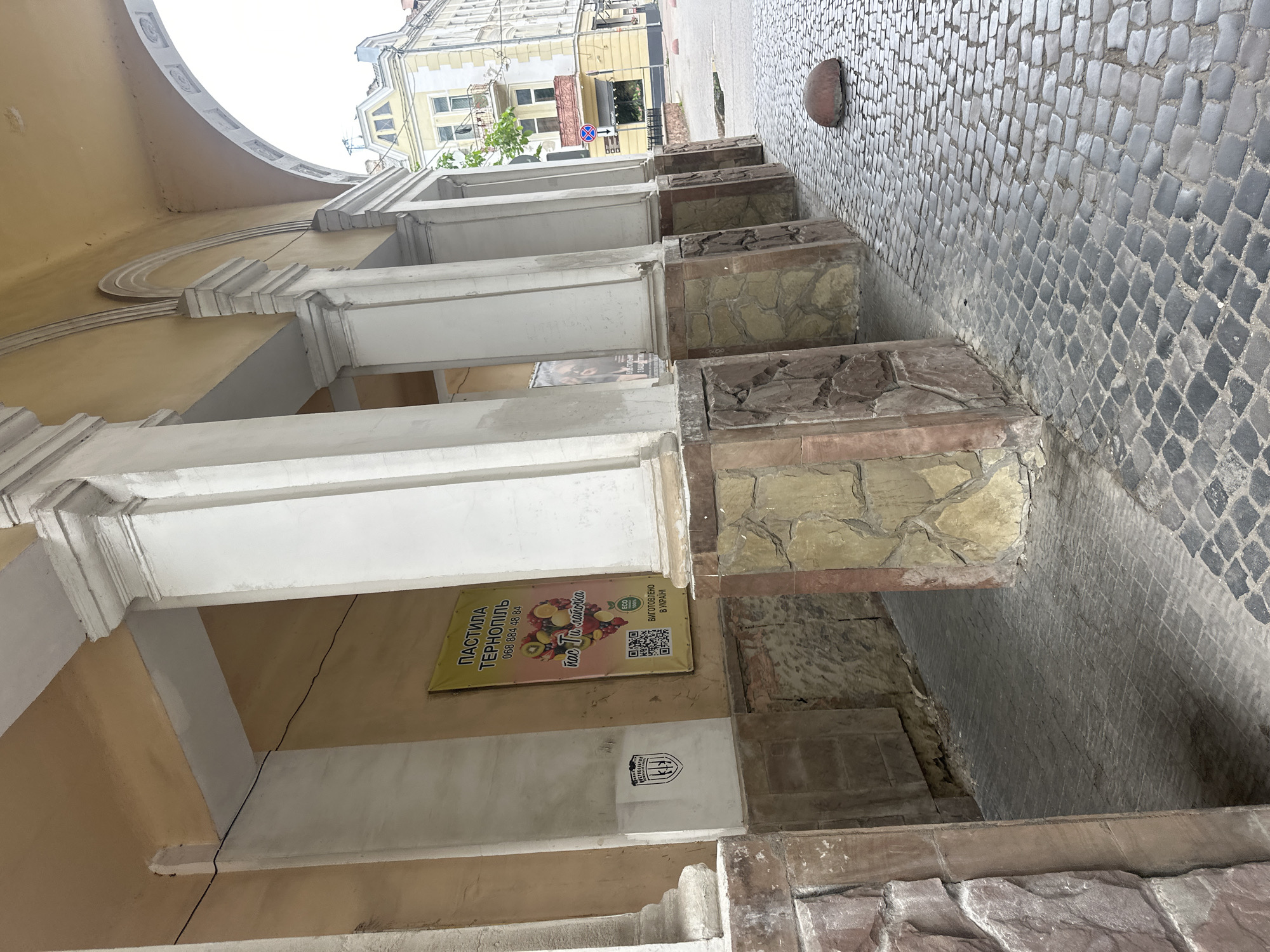 The image depicts an outdoor scene within a stone and plaster arched colonnade in Ternopil, Ukraine. The structure features multiple white classical pillars with capitals, supporting an overhead archway. The upper walls and ceiling of the passageway are painted a light ochre, while the lower sections of the pillars and wall bases are clad with rough-hewn stone in shades of brown, beige, and reddish-brown.
On the left wall, between two pillars, a yellow banner is displayed. It reads "ПАСТИЛА ТЕРНОПІЛЬ" (Pastila Ternopil) with the phone number "068 884 48 84" and the phrase "нас ти лапочка". A heart-shaped arrangement of various fruits (kiwi, lemon, cherries, apples, strawberries, raspberries, oranges) is centrally featured. Below this, two QR codes are visible, accompanied by "ECO 100%" and "ВИГОТОВЛЕНО В УКРАЇНІ" (Made in Ukraine). To the left of the banner, a black and white shield-shaped emblem, bearing the text "УКРБУДРЕНОВАЦІЯ" (UkrBudRenovatsiya), is affixed to the wall. A black electrical cable runs horizontally across the upper wall section.
The ground is paved with dark grey cobblestones, which extend through and beyond the passageway. To the right, on the cobblestones, a single reddish-brown, dome-shaped street bollard is present.
Through the archway, an exterior street scene is visible under an overcast sky. A light yellow building with white trim, multiple windows, and a distinctive gable roofline is prominent in the background. A smaller building with a dark storefront is partially visible to its right. A blue circular street sign with a red diagonal line (indicating "no stopping") is mounted on a pole. Green foliage from a tree or bush is also visible. No individuals are present in the scene.
