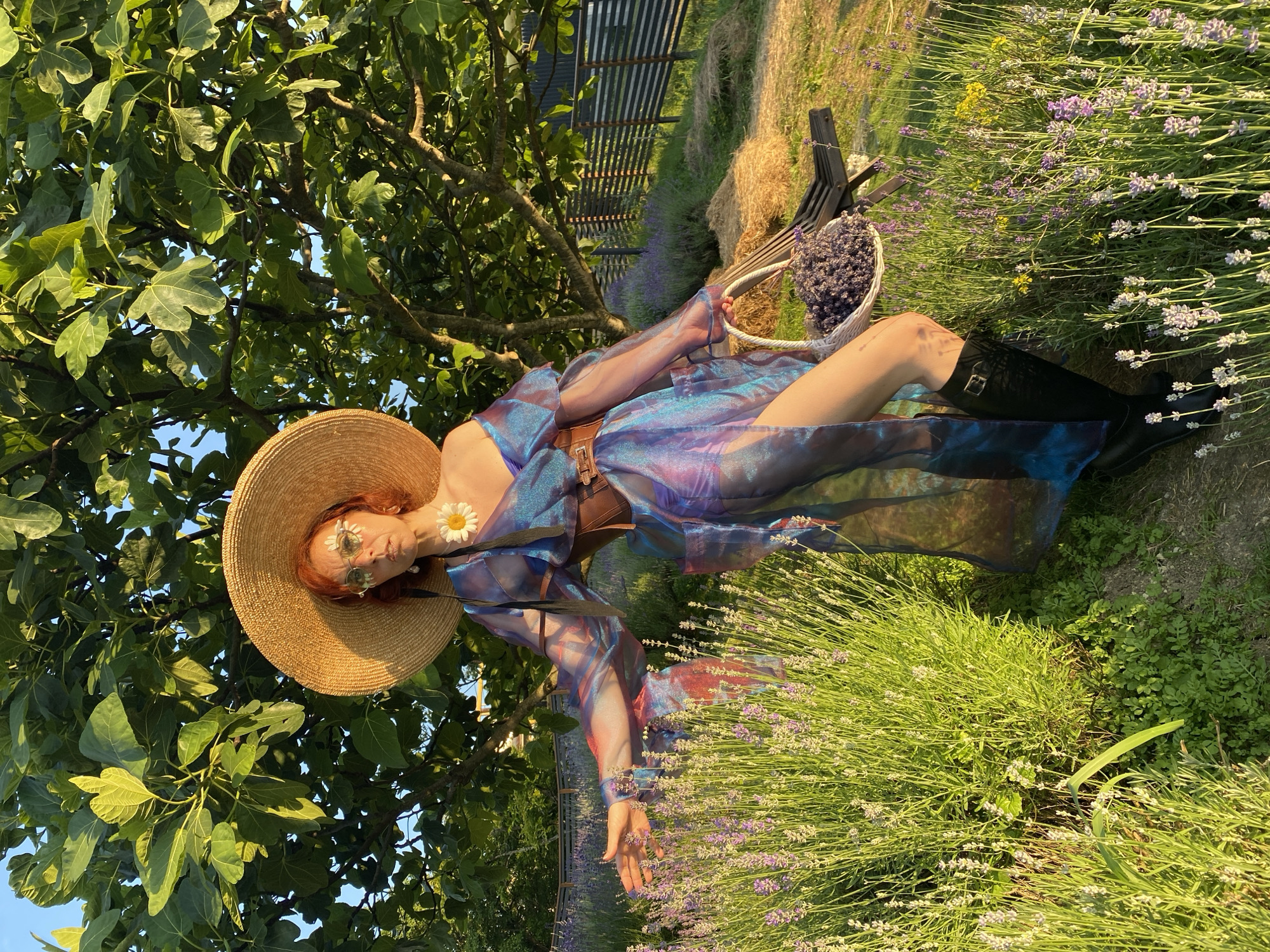 A person wearing a wide-brimmed straw hat, round sunglasses with daisy accents, and a sheer iridescent blue dress with a brown belt stands in a field of lavender. The person has auburn hair and is decorated with a daisy on their chest. They are holding a basket filled with lavender flowers. To the right, a dark wooden chair and hay bales are visible. The background features a large tree with broad leaves and a wooden fence. The scene is bathed in warm, golden sunlight, suggesting it is late afternoon or early evening. The location is the city of Verkhneye Uchdere, Russia.