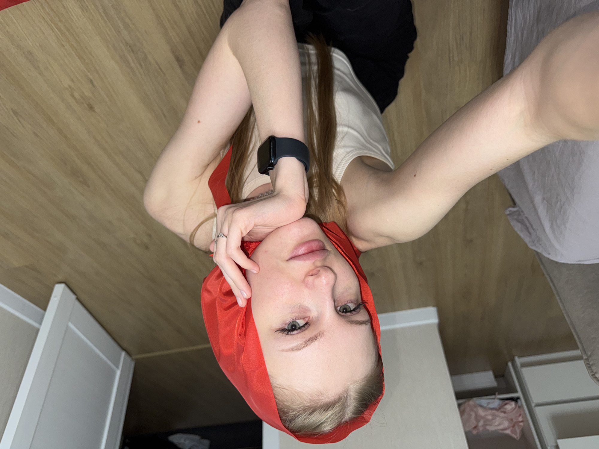 A young woman with blonde hair, wearing a red headscarf, a white tank top, and black pants, is pictured from the chest up. She is holding her chin with her right hand, revealing a ring on her right index finger and a black smartwatch on her left wrist. The background consists of a wooden floor, a beige wall, a white door, and a closet with some clothing items. The scene appears to be indoors, in a domestic setting. There is no information in the image to suggest a specific location in Perm, Russia.