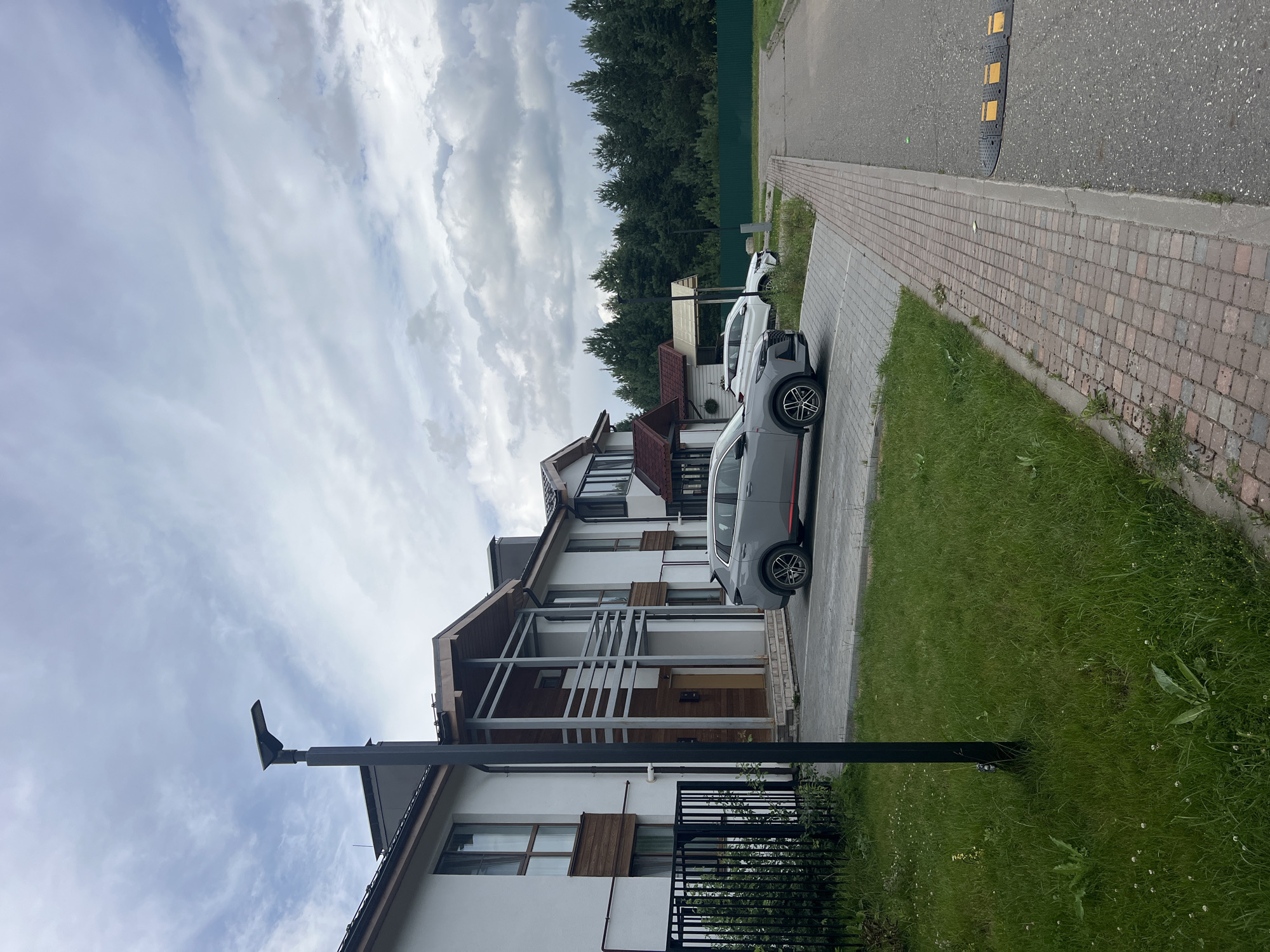 This is a medium-shot, exterior photograph taken on a cloudy day in Русский Вожой, Russia. The scene depicts a row of modern townhouses with white facades and wooden accents. In the foreground, a paved driveway with a speed bump is visible. Two cars are parked on the driveway: a gray SUV with a red stripe on the side and a white sedan behind it. A black lamppost stands in front of the townhouses. A black metal fence with green bushes borders the property. The background features a dense forest of green coniferous trees. The sky is overcast with a mix of light and dark clouds.