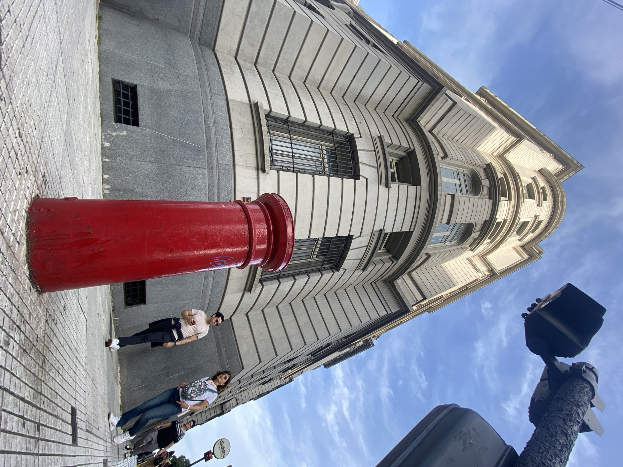 The image shows a low-angle view of a street scene in Buenos Aires, Argentina, featuring a red British-style post box. A tall, light-colored building with a curved facade and repeating horizontal architectural details dominates the background. Windows are visible with black bars. Two people are seen standing on the sidewalk next to the post box, another group of people behind them. The sky is blue with light clouds. A traffic light post with visible traffic lights is on the right side of the image. A "Subte" sign is at the bottom left corner. The sidewalk is made of light-colored paving stones.