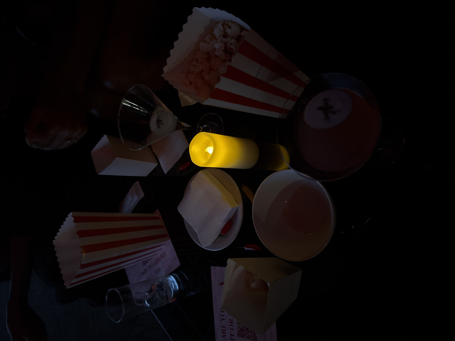 The image depicts an arrangement of cinema-related items scattered on a dark surface. A vibrant yellow cylindrical candle casts light upon a martini glass, popcorn boxes, and cups containing liquids. Discarded napkins and tickets are also visible. The scene suggests a post-movie setting, with the remnants of snacks and drinks. A dark pool ball can be seen on the upper right.