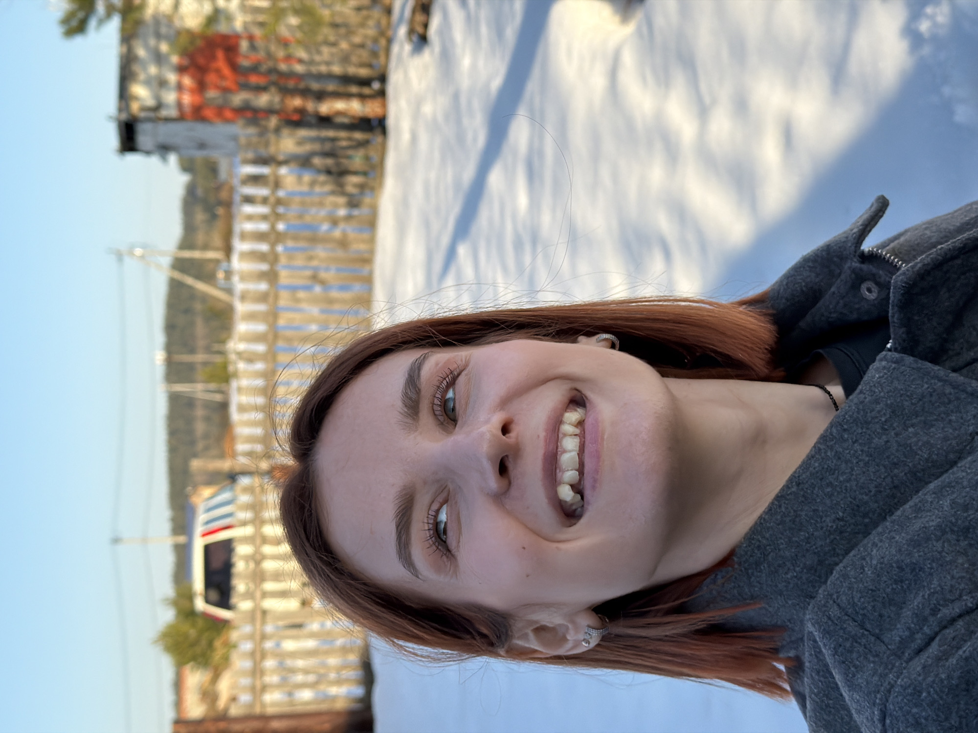 A young woman with auburn hair and fair skin smiles at the camera. She wears a gray jacket and a black top. The background shows a winter scene. In the distant background is a white fence and a building with red accents. Snow covers the ground. The location is possibly Chaikovsky, Russia based on user context.