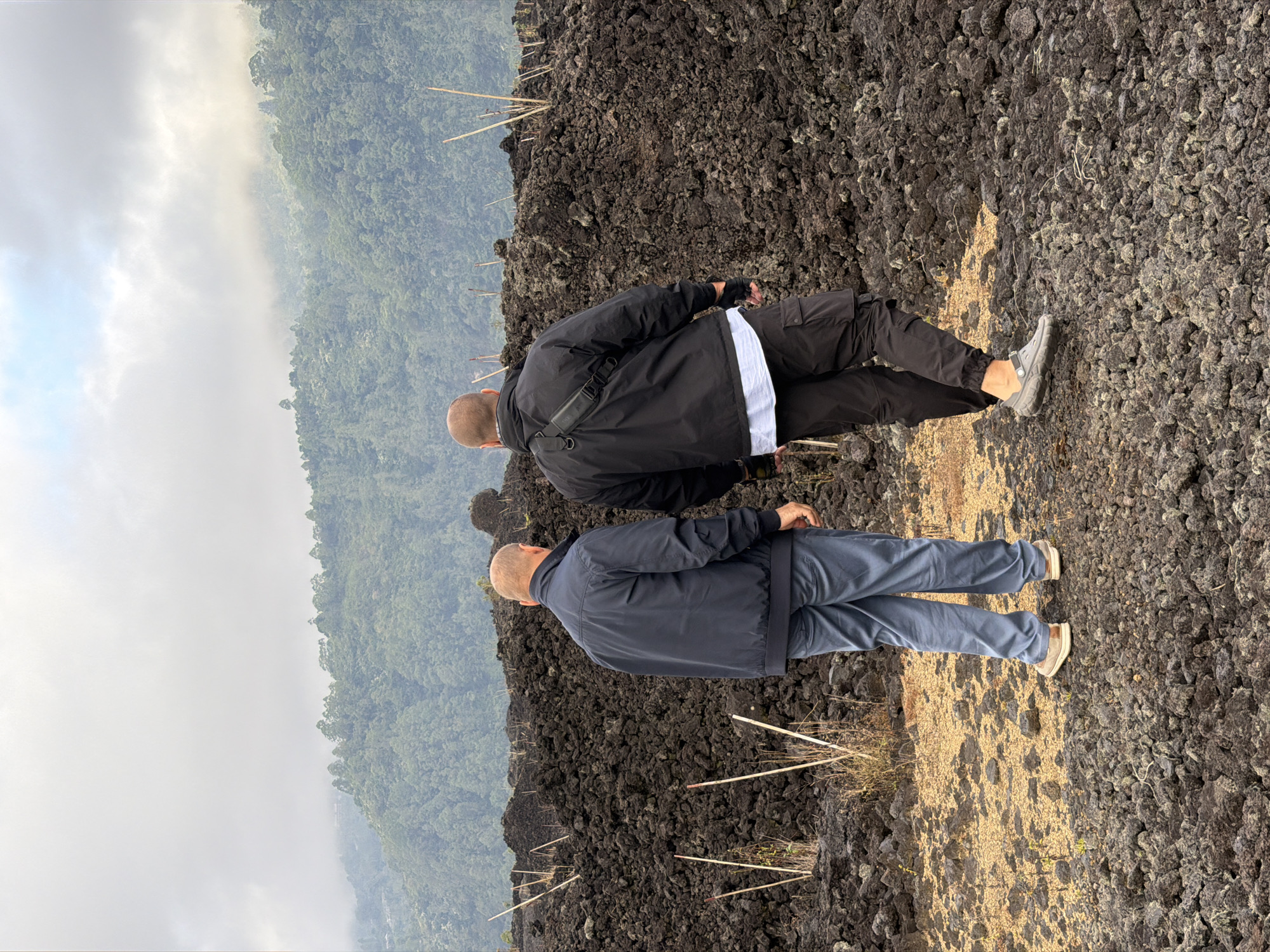 Two bald men are standing in a field of volcanic rock in Kintamani, Indonesia. The man on the left is wearing a dark blue jacket and grey pants. The man on the right is wearing a black jacket, black pants, and grey shoes. In the background, there is a lush green mountain covered in trees, with a cloudy sky above. The volcanic rock field is a mix of dark grey and black rocks, with some dry grass and thin wooden sticks scattered throughout. The scene appears to be a natural landscape, possibly a hiking trail or an area affected by a volcanic eruption.