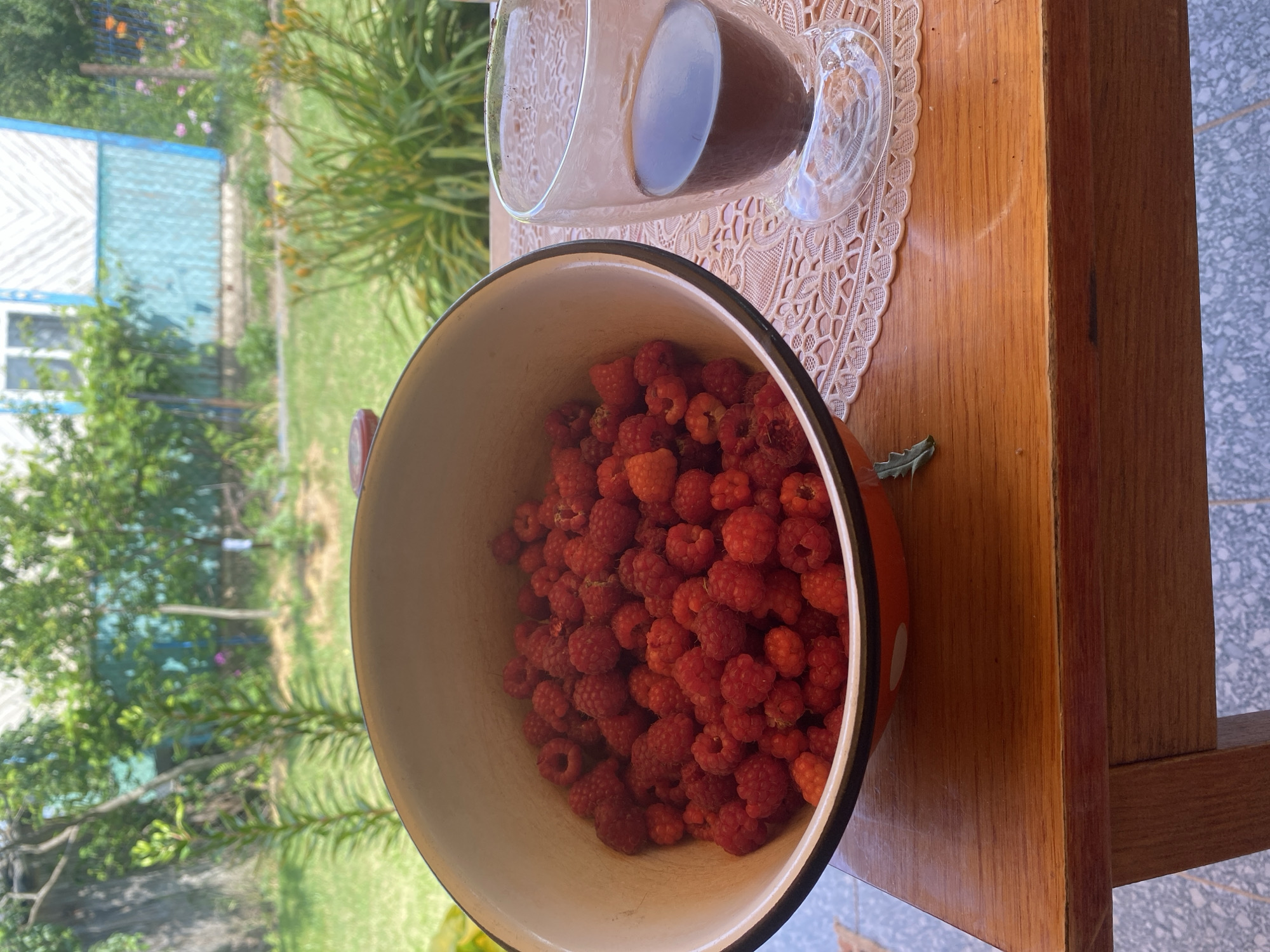 This image shows a close-up view of a bowl filled with ripe raspberries, resting on a wooden table outdoors. To the right of the bowl, a clear glass mug containing a dark liquid, likely coffee or tea, is positioned on a white lace doily. The background is blurred but suggests a garden setting with green foliage and a hint of a blue and white building, possibly a house or shed. The scene appears to be set during the daytime, with natural light illuminating the table and the raspberries. The overall impression is one of a simple, pleasant moment, perhaps a break for a snack or refreshment in a rural or suburban environment. The focus is on the fresh fruit and the drinks, suggesting a moment of enjoyment. There is no visible text in the image.