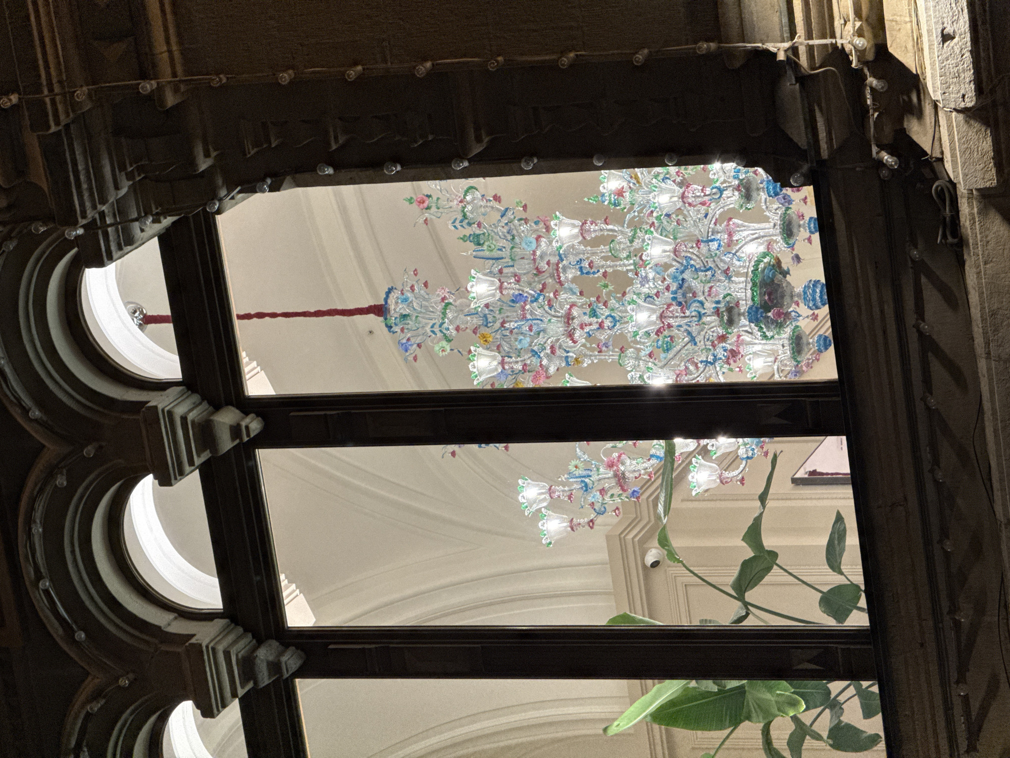 The image shows a view through a decorative window with an arched design, likely from inside a building. The window frames are dark and detailed. Through the window, the focus is on a large, ornate chandelier with many glass elements, possibly made of Murano glass, adorned with floral and colorful accents. Part of the interior ceiling is visible, with arches and architectural details. Additionally, a plant with broad green leaves can be seen partially obstructing the view through the lower part of the window. The composition suggests the photo was taken in an upscale interior setting, and based on the architecture and decoration, it could potentially be located in Moscow, Russia. A small white security camera is visible in the background.