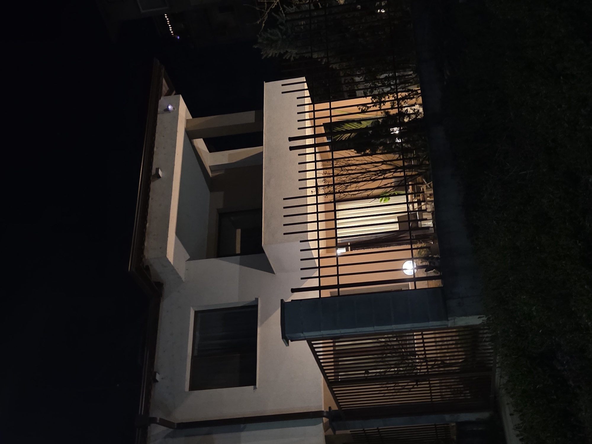 At night, a multi-story building is illuminated. The building has a modern architectural design with a concrete facade and multiple balconies enclosed by horizontal slats. A large dark window is visible on the lower level. The lighting inside reveals hints of interior features, including what appears to be furniture and a circular light fixture. Dense vegetation is present to the right of the building. The dark sky suggests the photo was taken late in the evening or at night. I do not have information to verify the location.