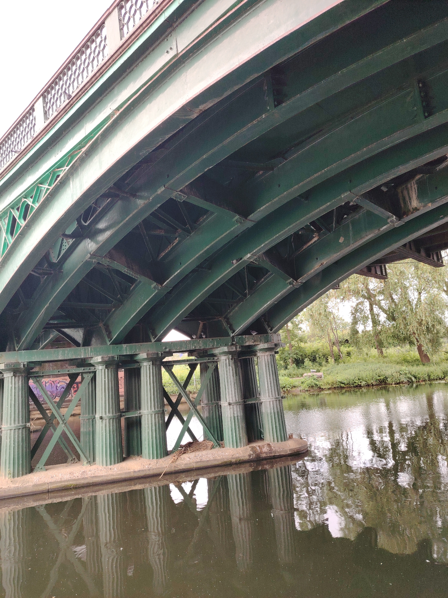 The image depicts the underside and a portion of the top railing of a large, dark green metal bridge spanning a calm body of water, likely a river or canal, in Peterborough, United Kingdom. The bridge features an arched structure with complex crisscrossing metal beams, girders, and internal latticework visible from below, secured by numerous rivets. The upper edge of the bridge, visible in the top left, features a decorative, lighter-colored railing with repeating circular or ovoid patterns.
The bridge is supported by a series of robust, fluted or ribbed cylindrical metal columns painted the same dark green, which rise from a concrete or stone base directly in the water. Between these columns, diagonal metal cross-bracing forms an 'X' pattern. Graffiti in purple and white spray paint is visible on the concrete base and lower sections of the columns.
The water below the bridge is dark, brownish-green, showing reflections of the bridge structure, supports, and the trees lining the far bank. Small ripples disturb the water surface, and minor floating debris, including twigs and leaves, is scattered on the water, with a small accumulation of sticks visible at the waterline of a bridge support on the right.
The far bank, visible to the right and through the bridge supports, is densely vegetated with various green trees, including a prominent willow-like tree with drooping branches, bushes, and grasses, under a bright, overcast sky. A small, grey bird is perched on a structural element of a bridge support on the left side of the image, near the waterline. No people are present in the scene.