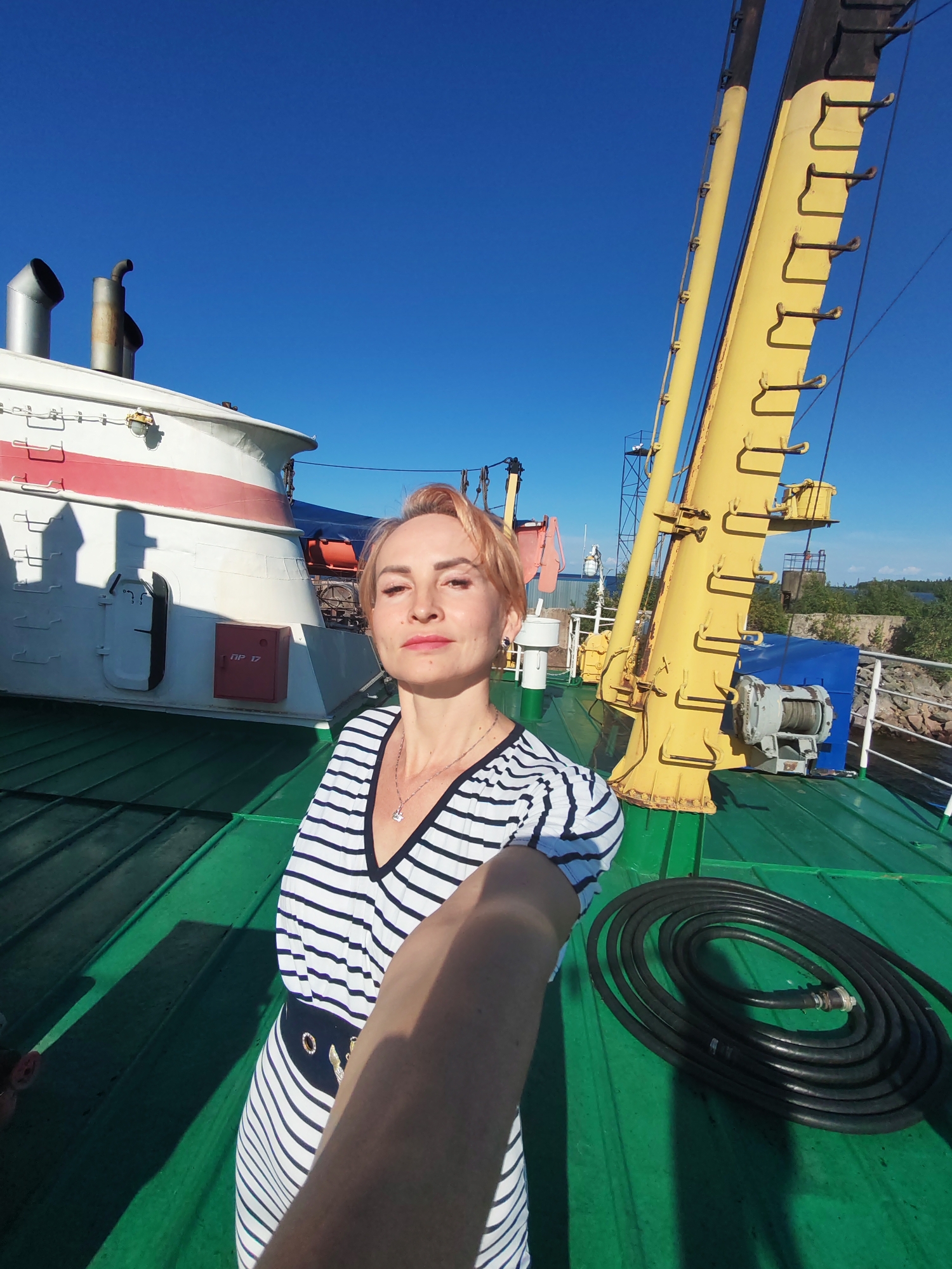 The image shows a woman taking a selfie on the deck of a ship. She is wearing a black and white striped dress and a necklace. Behind her is a yellow crane and part of the ship's superstructure, which includes smokestacks and a red box. The deck is painted green, and a coiled black hose is visible on the deck. The sky is clear and blue. The location is the port of Kvartsitny, Russia.