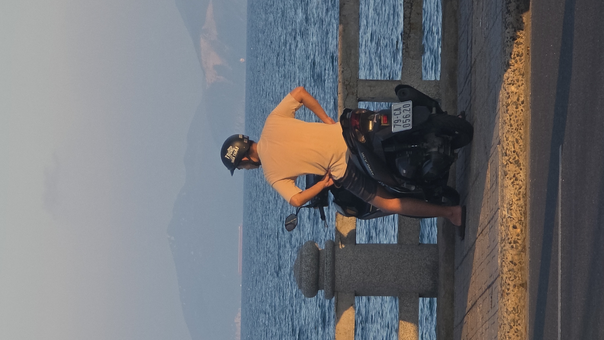 The image depicts a single male, positioned on a black scooter, parked on a paved sidewalk adjacent to a concrete sea barrier. The scene is located along a coastal road in Nha Trang, Vietnam, indicated by the scooter's license plate, 79-CA 056.20, which corresponds to Khanh Hoa province.
The man, viewed from behind, wears a light-colored, short-sleeved t-shirt, dark shorts, and dark flip-flops. A black helmet with white text is worn on his head. His hands are placed on his hips, and his posture indicates he is observing the ocean. The scooter, identifiable as a Honda Air Blade, has a black exhaust system.
In the mid-ground, the barrier comprises grey concrete posts and horizontal rails separating the sidewalk from the sea. The ocean exhibits a blue hue with subtle surface ripples. A substantial, dark cargo ship is visible far on the horizon, with a smaller vessel to its right. The distant background features hazy, blue-grey mountains rising from the sea. The sky above is a pale grey-blue with minimal cloud cover.
The foreground consists of a light-colored, textured pavement, bordered by a rough-textured concrete curb. Beyond the curb, a dark asphalt road with a faint white dividing line is visible. The lighting suggests a late afternoon or early evening setting, with warm tones highlighting the man's back and the foreground pavement, and casting long shadows across the road.