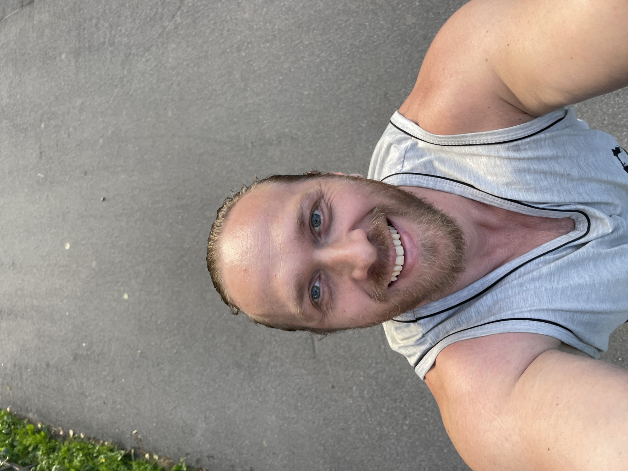 The image is a selfie taken from a high angle looking down at a man with light skin, blue eyes, and a light brown beard. He has wet, light brown hair pulled back from his forehead. He is wearing a light gray sleeveless tank top with black trim. The background is an asphalt path with a grassy area on the left side of the frame. The scene is outdoors, with natural lighting. It does not appear to be in Saint Petersburg, Russia.