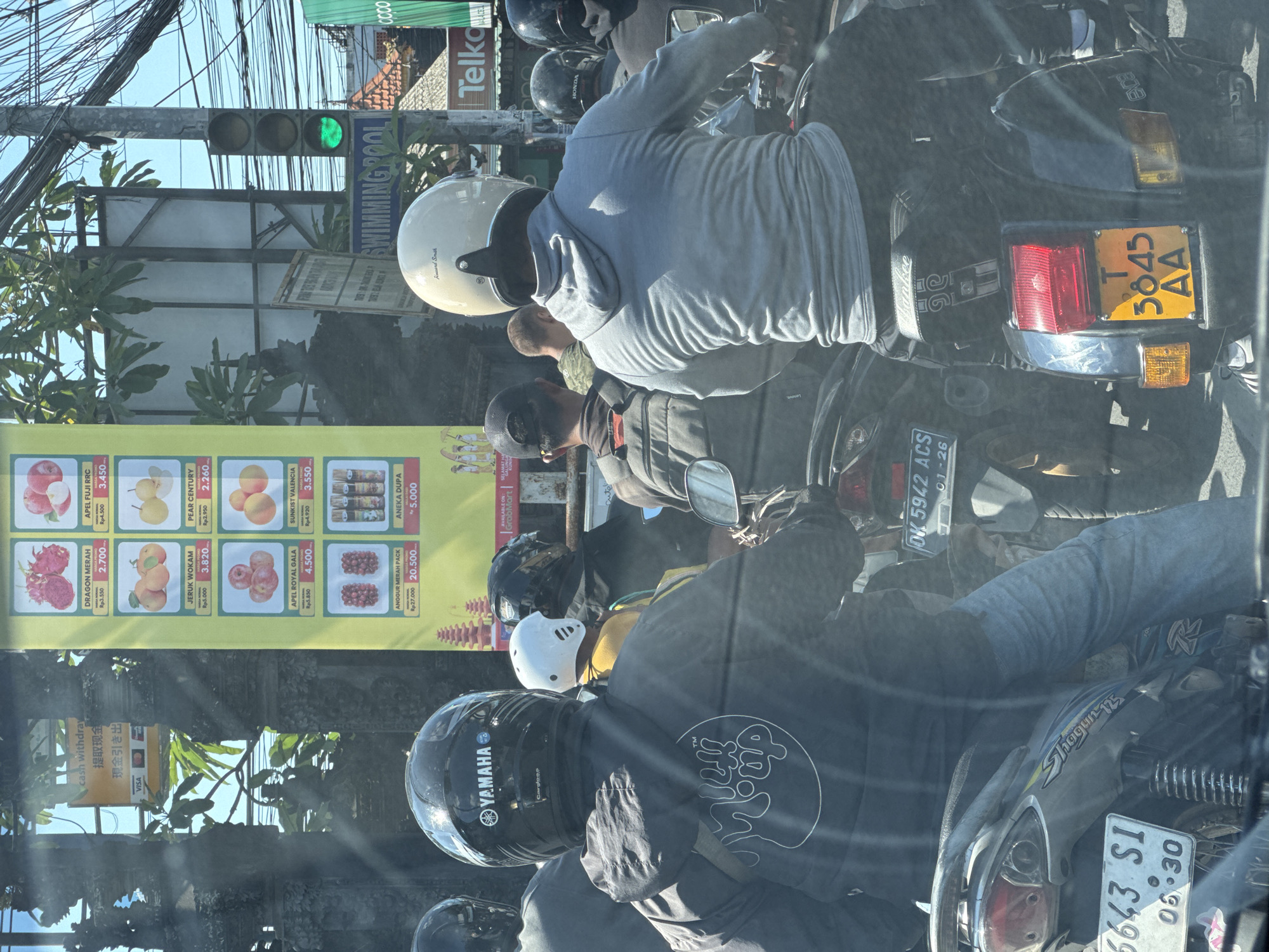 The image captures a street scene in Kuta Selatan, Indonesia, likely during daylight hours given the strong sunlight and shadows. The foreground is dominated by the backs of several individuals on motorcycles, suggesting a traffic situation. A prominent black helmet with a "YAMAHA" logo is visible, as is a light-colored helmet with the inscription "Howard Smith". The individuals are wearing jackets and casual clothing.
The environment includes street infrastructure: a traffic light displaying a green signal, power lines, and street signs. One sign reads "SWIMMING POOL". To the left, a vertical yellow banner displays images and prices of fruits, including "DRAGON MERAH", "APEL FIJI RRC", "JERUK WOKAM", and "PEAR CENTURY". Another banner features advertisements for "ANEKA DUPA".
The motorcycles themselves are a significant element. One partially visible motorcycle in the lower left has a license plate reading "6643 S1 06.30". Another motorcycle in the middle ground has a license plate "DK 5942 ACS 01-26". A scooter in the right foreground has a yellow license plate with the characters "T 3645 AA". The overall atmosphere appears to be that of a busy, possibly congested, urban or semi-urban area.