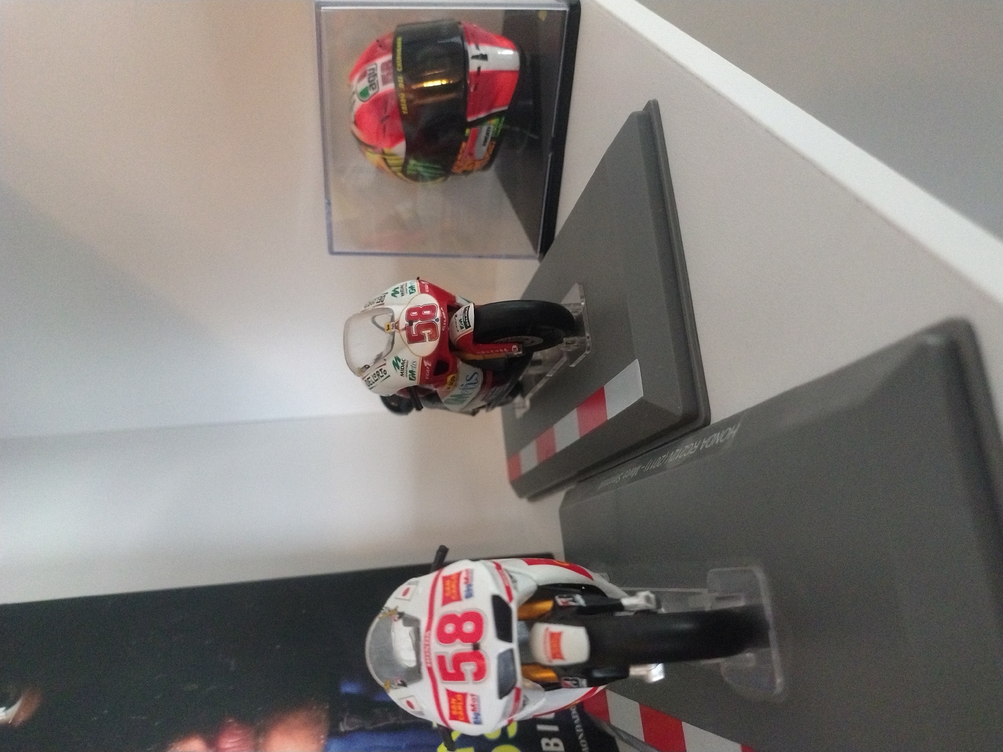 The image shows a display of motorcycle memorabilia, likely miniature models, arranged on a white shelf or wall. The items include two miniature motorcycle models mounted on gray stands with red and white striped sections, and a helmet in a clear display case. The number "58" is visible on both motorcycles. The photograph appears to be taken at an angle, giving a slightly tilted perspective of the display. Text is visible on the stands and possibly on the motorcycle models, including brands like "DELLORTO", "MIDAC", "Getis", and the motorcycle model "HONDA RC212V (2011)". The location in the city of Crauthem, Luxembourg is not discernable from the image alone.