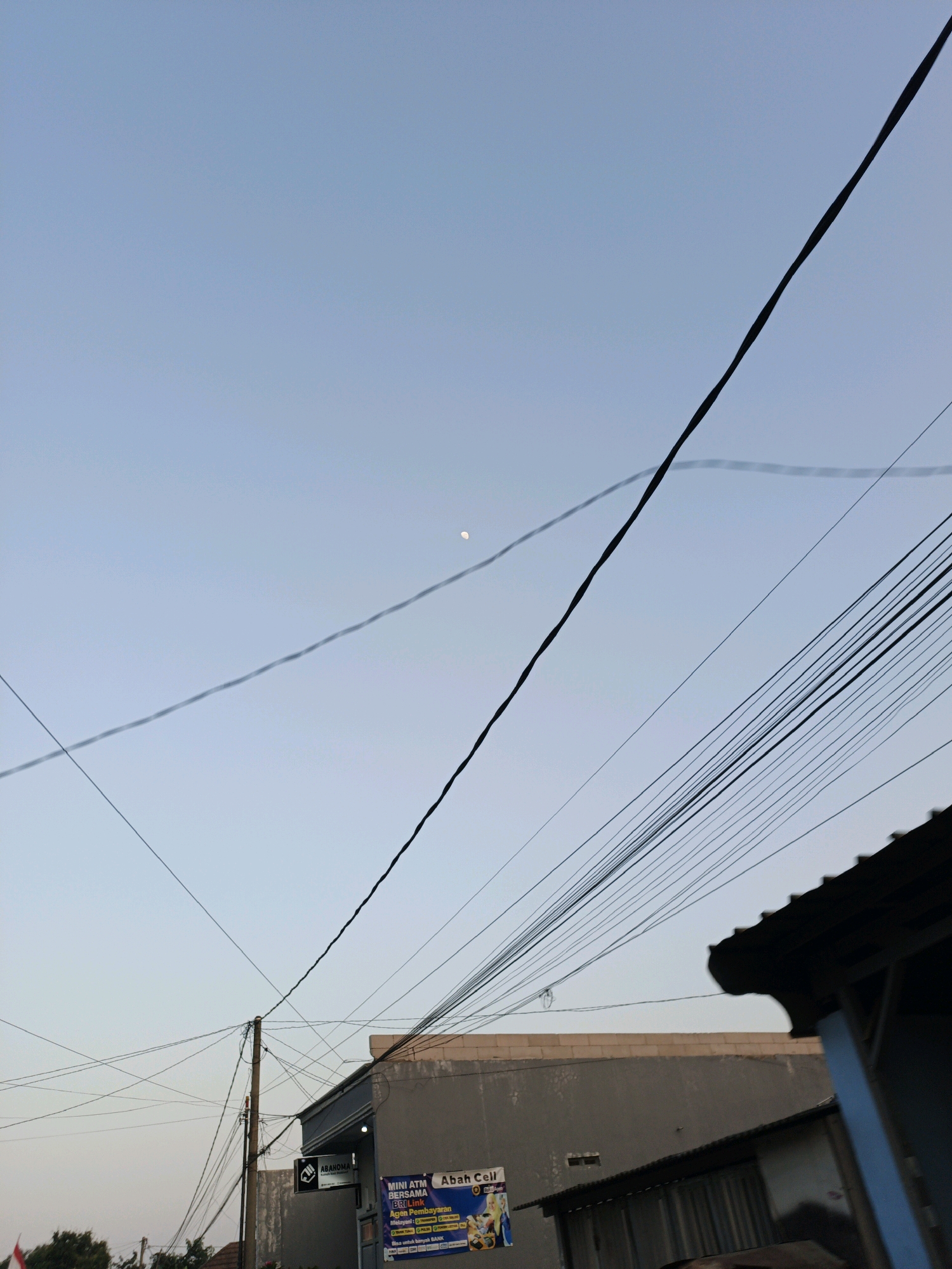 The image depicts a daytime sky with scattered clouds and a sliver of the moon visible. A dense network of electrical wires crisscrosses the frame, casting faint shadows. In the foreground, the roof of a building, possibly a small shop or residence, is visible on the right. A sign on a nearby building reads "MINI ATM BERSAMA BRI-LINK Agen Pembayaran Abah Cell", indicating a financial service point. The overall scene suggests an urban or semi-urban environment in Muntur, Indonesia, characterized by utility infrastructure and local businesses. There are no people or animals visible in the image.