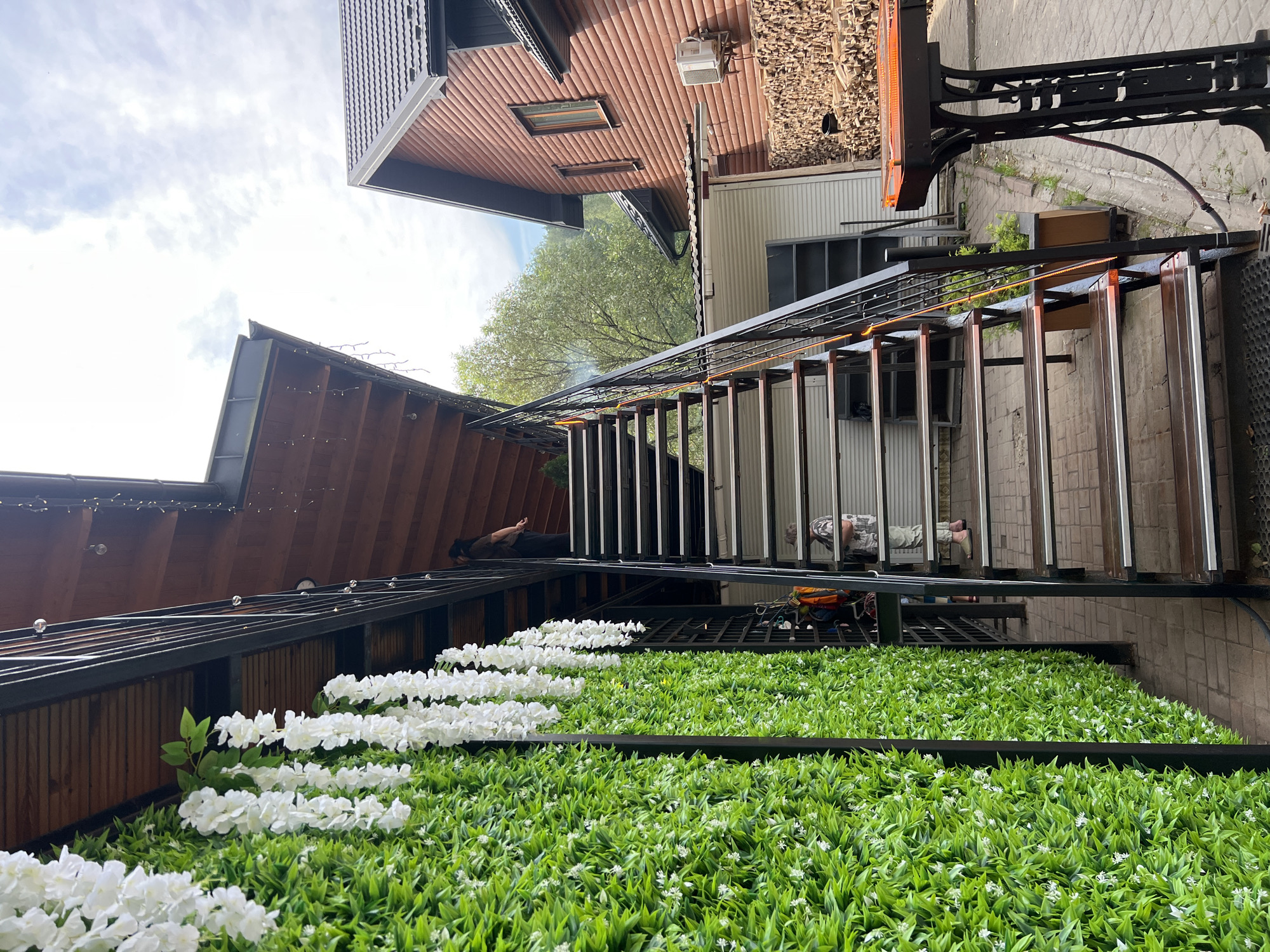 An outdoor staircase with wooden treads and metal railings leads up to a wooden-roofed structure. The left side of the staircase is adorned with a vibrant green artificial hedge wall featuring cascading white artificial flowers. On the staircase, two individuals are visible: one person stands near the top, looking away from the camera, and another person is on a lower step, facing the camera and appearing to be in casual attire. In the background, a wooden house with a dark roof and a pile of firewood stacked against its side is visible. The scene suggests a pleasant, possibly resort-like, outdoor setting. The image was taken in the city of Yaremche, Ukraine.