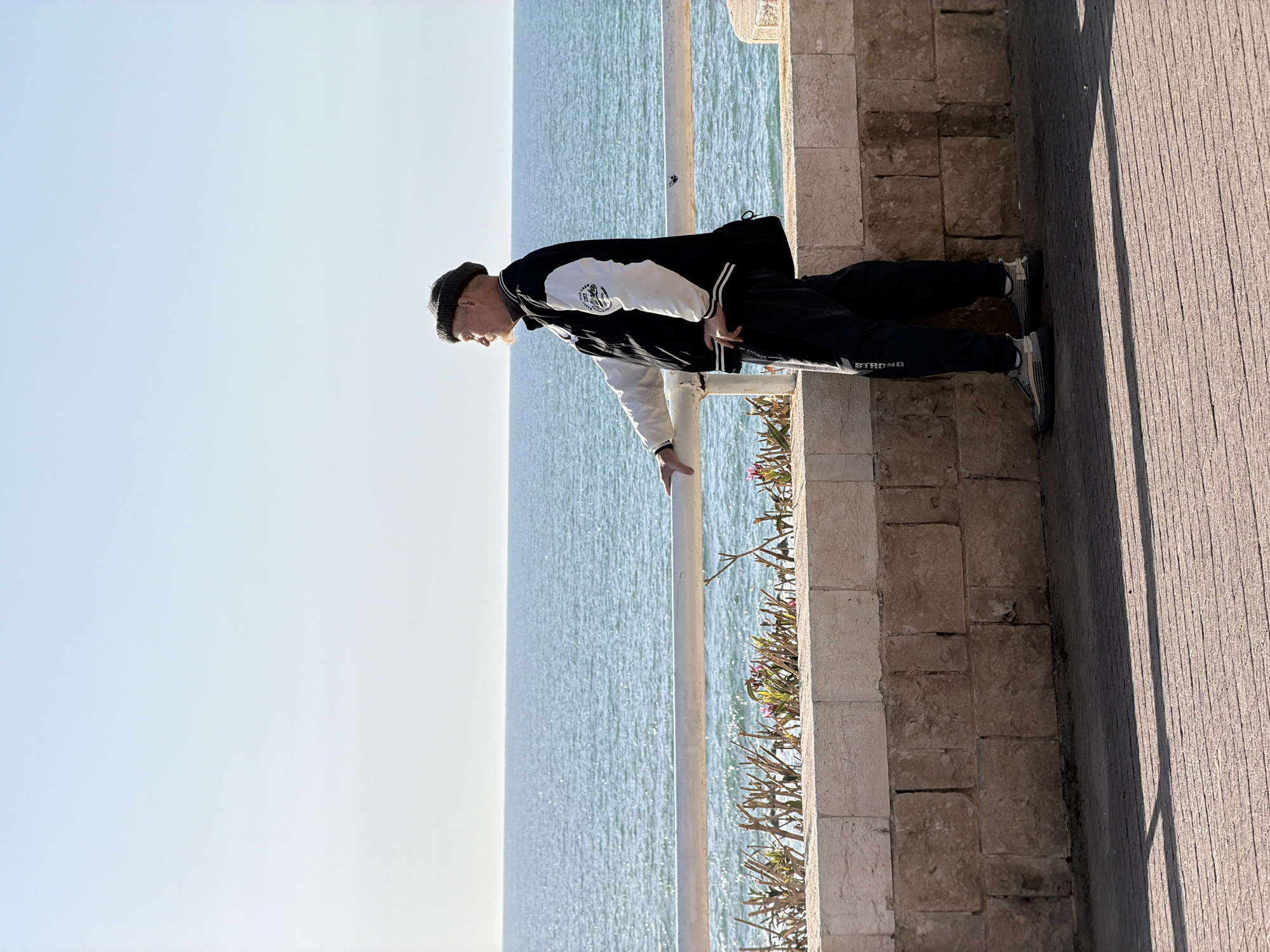 A man leans over a railing overlooking the sea. He wears a black beanie, a black and white jacket, and black sweatpants with the word "STRONG" down the side. His left hand is on the railing. The sea stretches to the horizon under a blue sky. The railing is next to a low stone wall, adjacent to a sidewalk. The location is in Bat Yam, Israel.