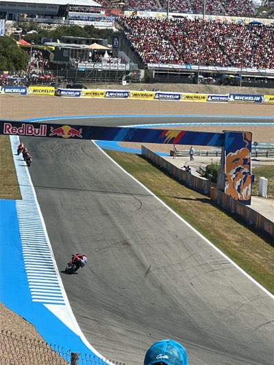 The image shows a view of the Circuito de Jerez-Ángel Nieto racetrack in Jerez de la Frontera, Spain. The track is visible, with two motorcycles racing on it. A Red Bull archway is present above the track. In the background, there are stands filled with spectators. Along the perimeter, there are "Michelin" banners. The environment is sunny. A blue cap is partially visible in the bottom center.
