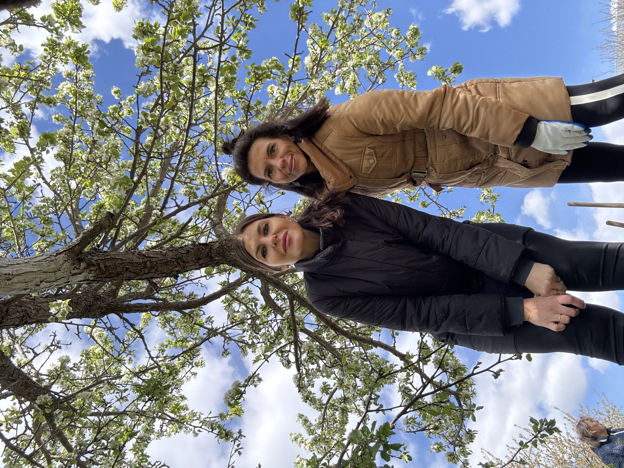 Two women stand under a tree with white blossoms against a blue sky with scattered white clouds. The woman at the top wears a tan jacket, black pants with a white stripe down the side, and a gardening glove on her right hand. The lower woman wears a black jacket and black pants, holding her hands together. The photograph is taken from a low angle, looking up through the tree's branches and blossoms. The location is the city of Dubovaya, Russia. An older person is partially visible at the bottom right corner of the image.