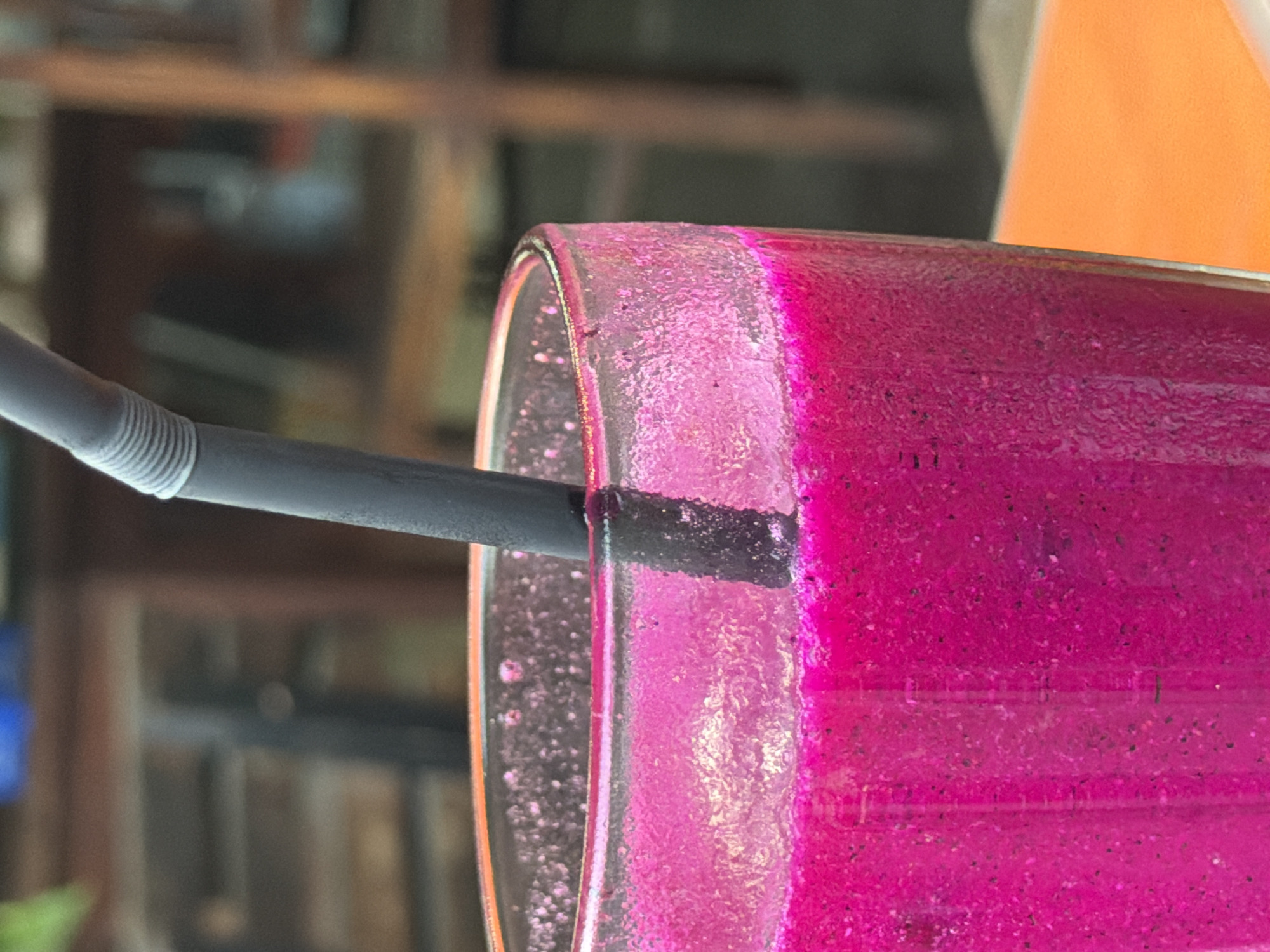 A close-up, shallow depth of field shot shows a glass of pink dragon fruit smoothie with a black straw. The glass is condensation-covered and filled to the brim. The smoothie has a textured appearance with small dark flecks visible. The background is blurred, showing wooden furniture and a hint of an orange surface in the foreground. The scene appears to be set at a cafe or restaurant in Kuta Selatan, Indonesia.