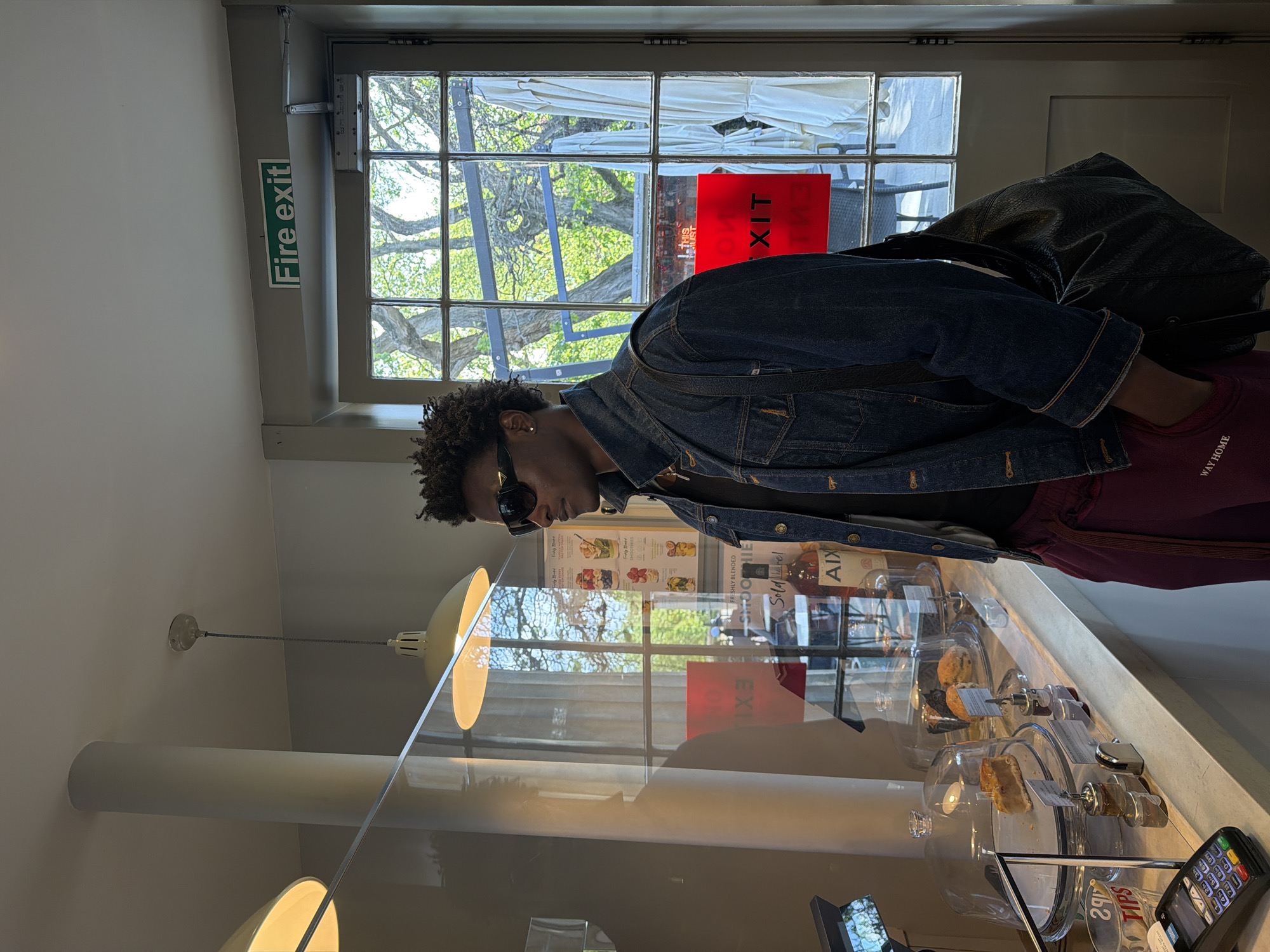 The image depicts an indoor scene within what appears to be a cafe or similar establishment, set during daytime in London, United Kingdom. A young person, characterized by their dark, curly hair and sunglasses, is the primary subject. They are wearing a dark denim jacket over a maroon top that has "WAY HOME" written on it, and a dark shoulder bag is visible resting on their back.
The person is positioned facing or looking down at a counter, suggesting they are in the process of ordering or browsing. The counter features a clear protective screen, behind which various baked goods, including muffins and cake slices, are displayed under glass cloches. Nearby, bottles labeled "AIX" and "Sol de" are visible. A poster advertises "SMOOTHIES FRESHLY BLENDED." A payment terminal showing "29" and "TIPS" can be seen on the counter.
A large window dominates the background, offering a bright view of the outdoors. Green trees with full foliage, indicating a sunny day, are prominent. White awnings or outdoor umbrellas are also visible through the window. On the interior wall above the window, a green sign clearly reads "Fire exit." A red sign with the text "EXIT" is partially visible through the window, likely an outdoor sign or part of an adjacent structure. The ceiling features light-colored pendant lamps. The overall atmosphere conveyed is casual and bright.